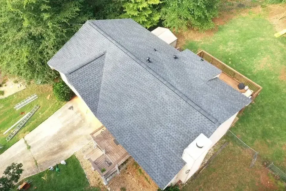 Aerial view of a house with a dark gray shingled roof, surrounded by green grass and trees.