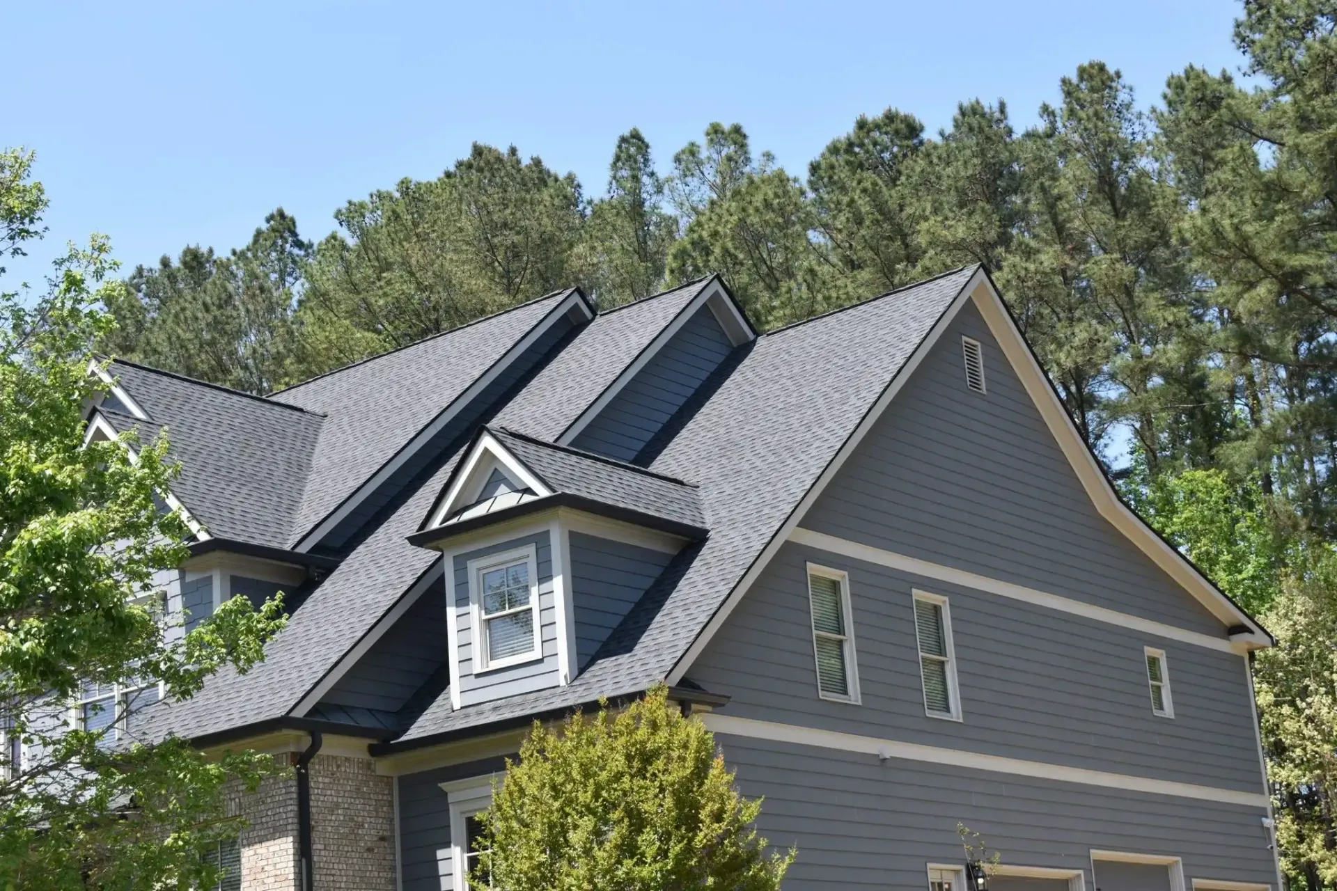 Gray house with dark gray roof and dormers, surrounded by green trees against a blue sky.