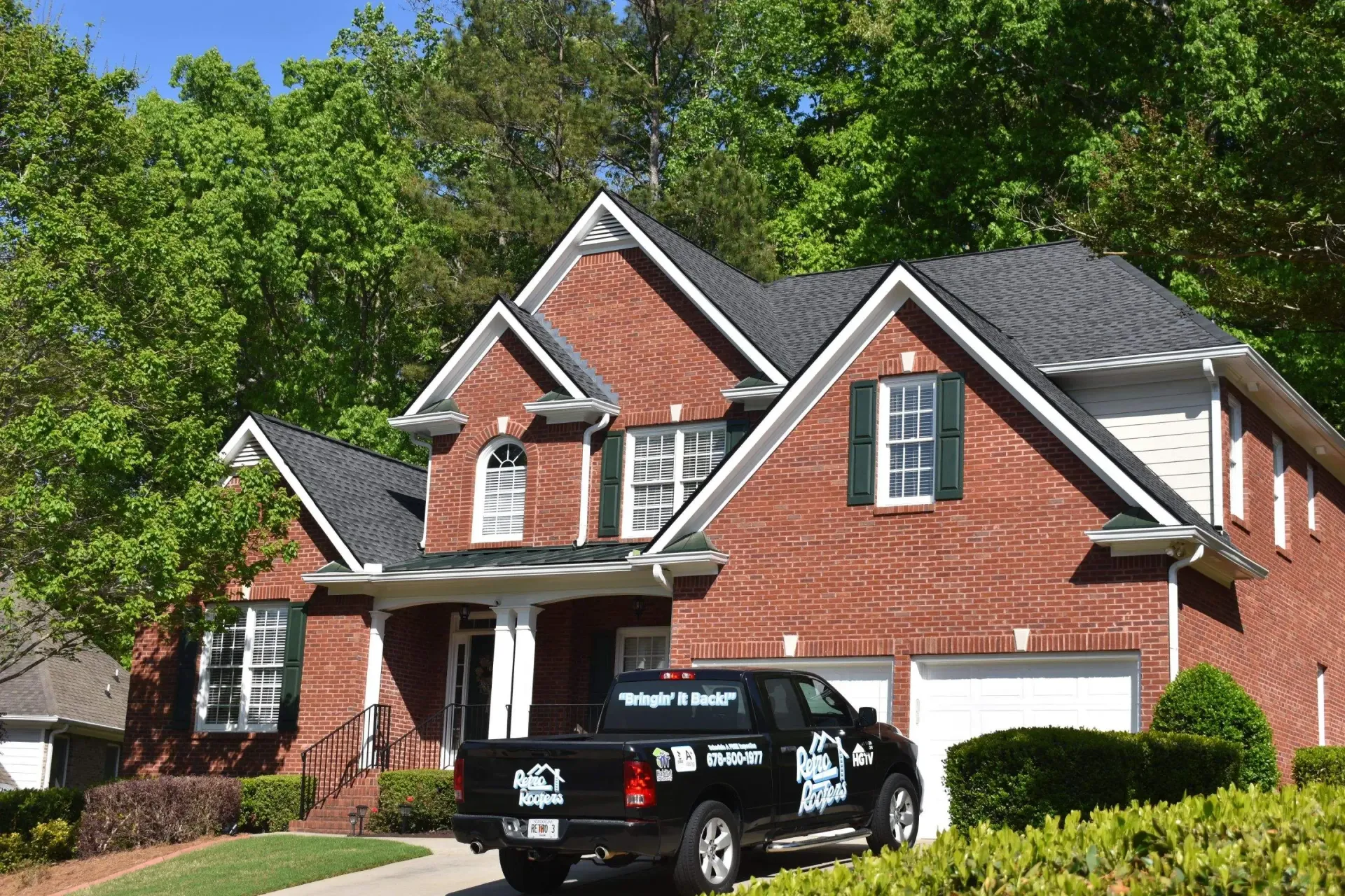 Brick house with dark roof, black truck parked in front of garage, green trees in background.