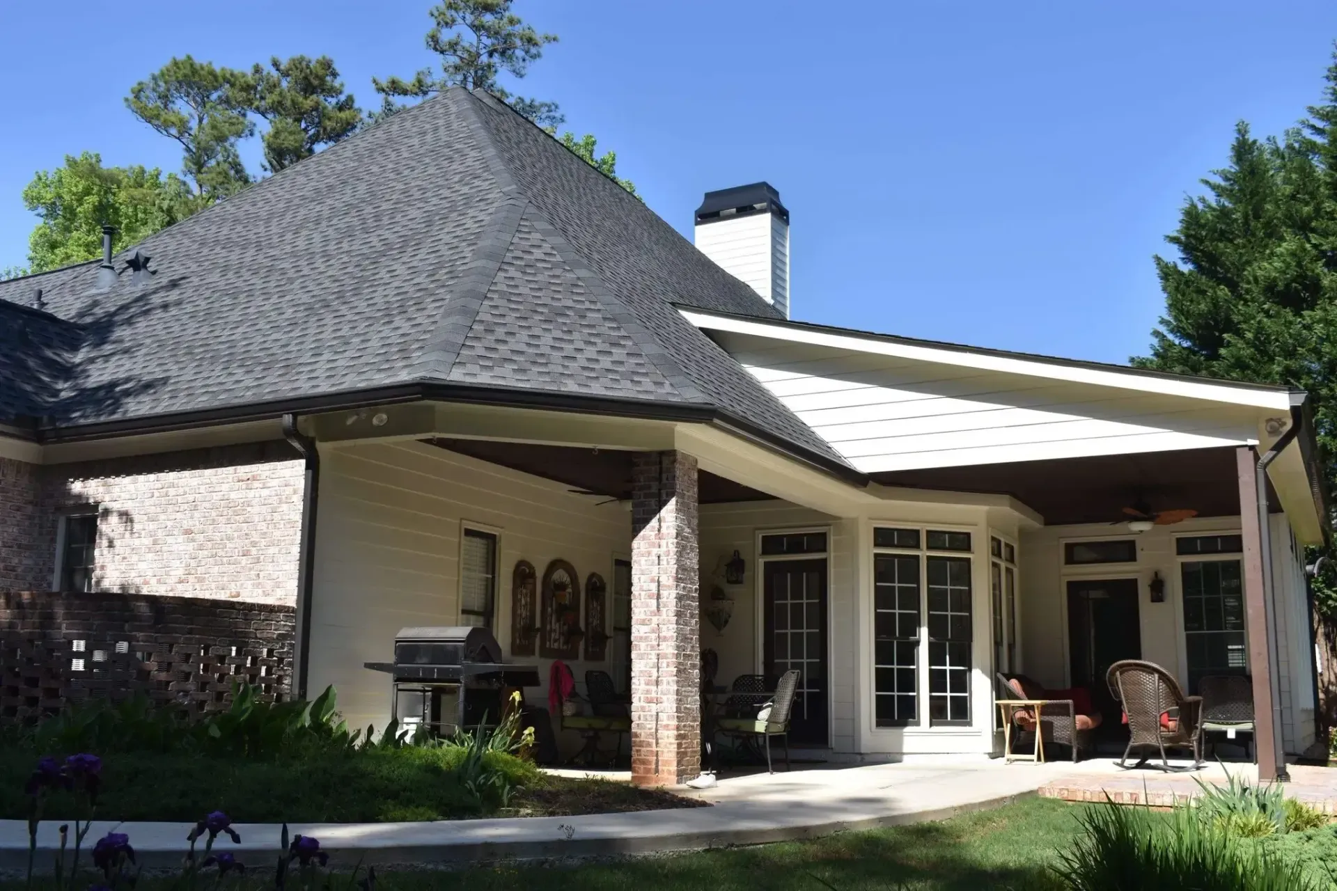 Tan house with gray roof and white trim, porch, and garden, under a blue sky.