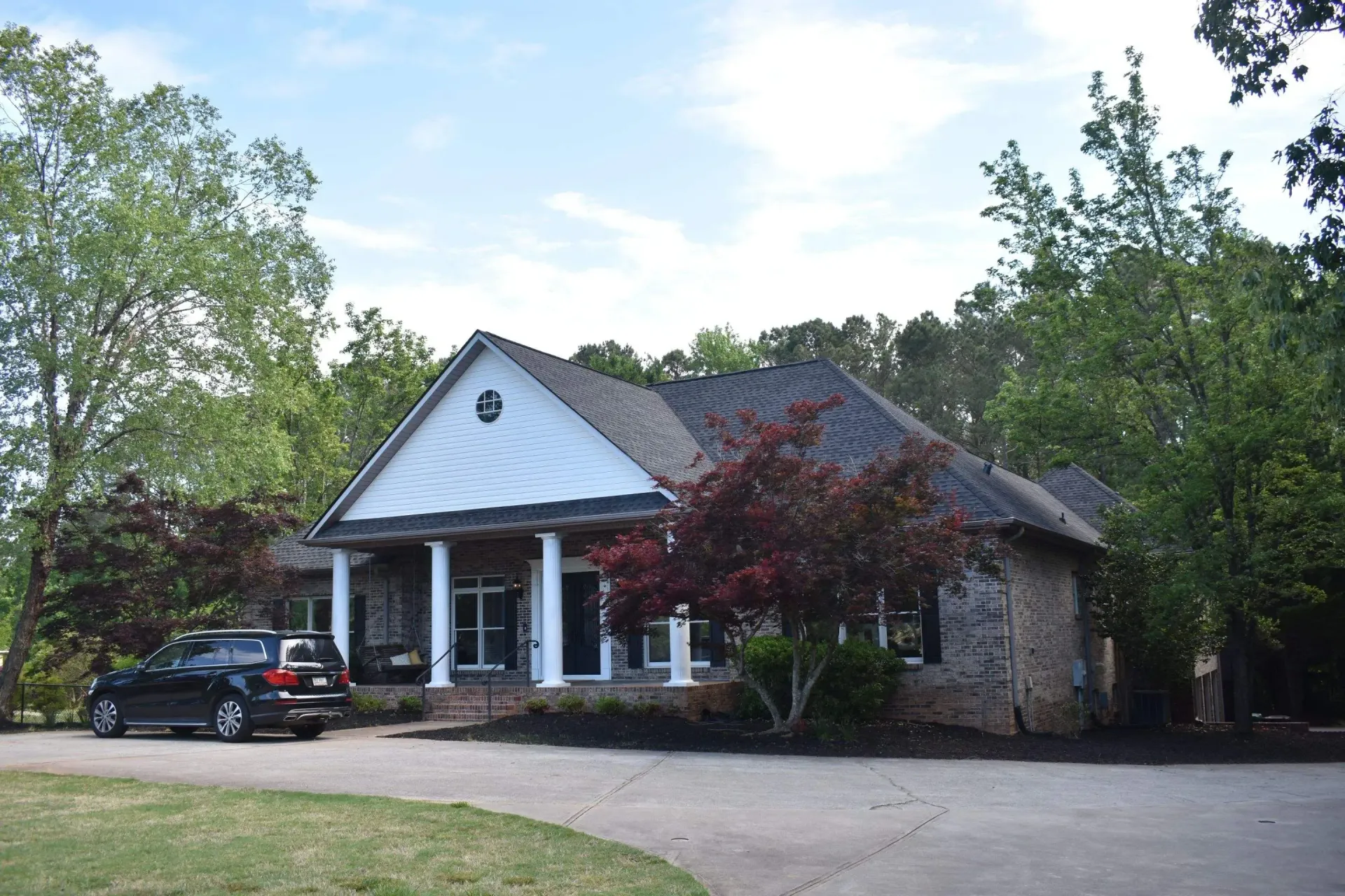 A one-story brick building with white pillars, a black car parked out front, surrounded by trees.