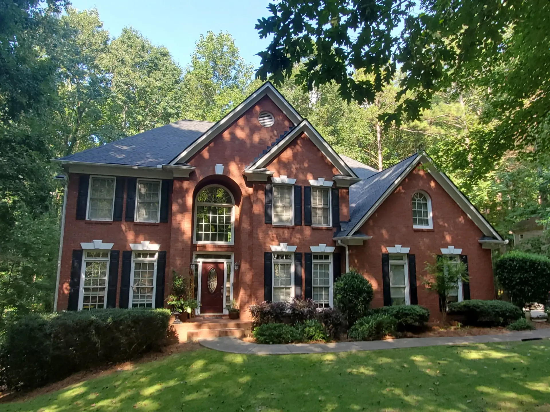 Two-story brick house with black shutters, red door, and lush green lawn in a wooded setting.