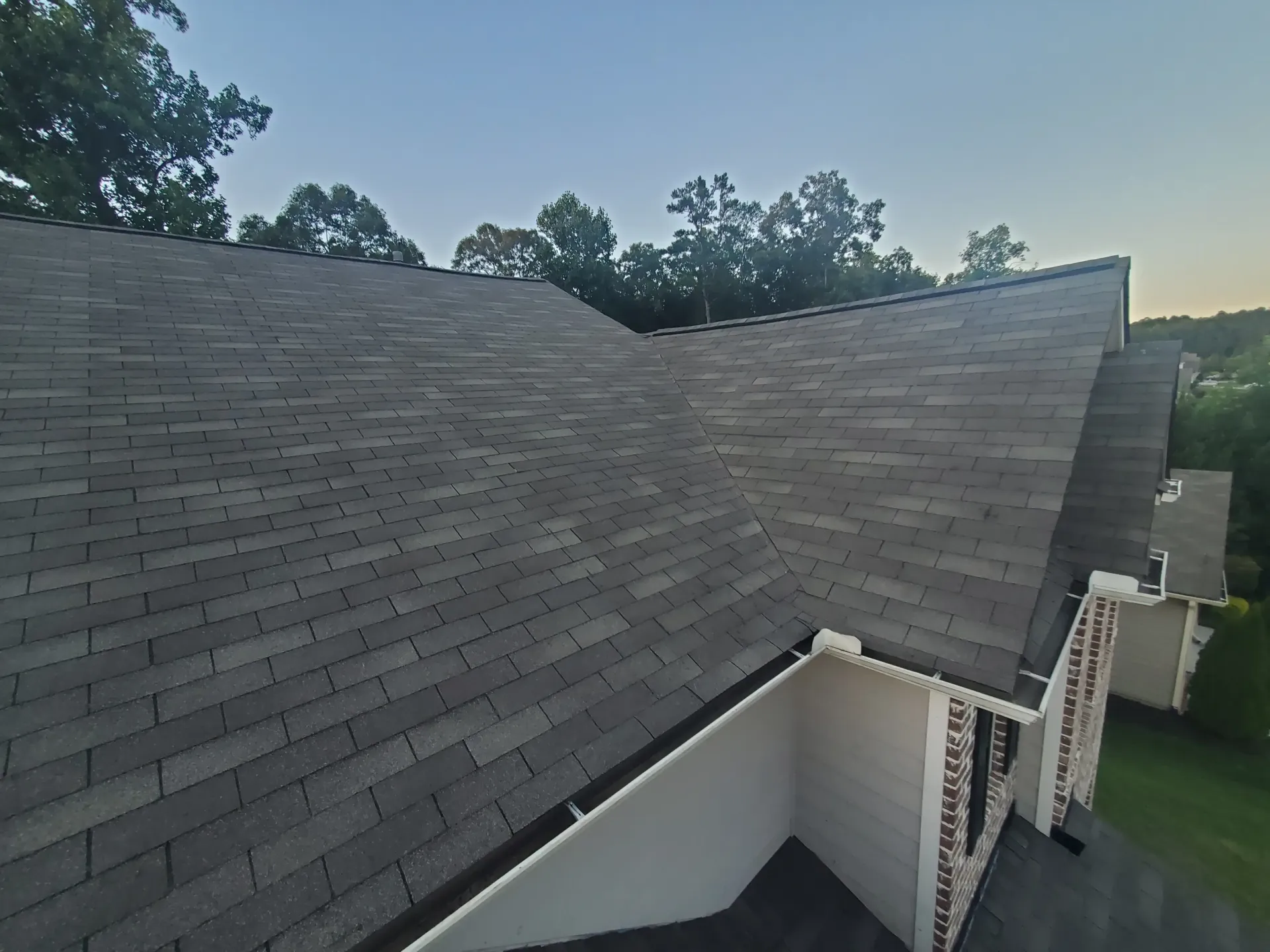 Gray asphalt shingle roof on a house with trees in the background under a blue sky.