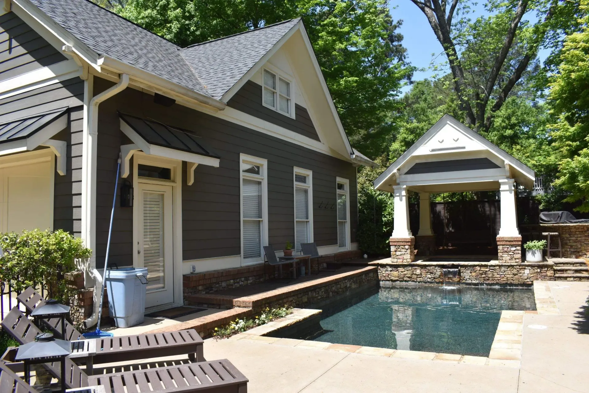 House with a dark exterior, pool, and gazebo. Blue pool water, lush green trees and sky.