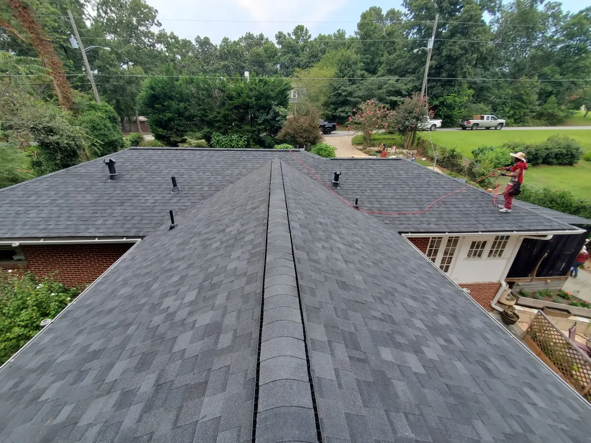 Dark asphalt shingle roof with black vents; a person stands on the right edge near trees and a street.