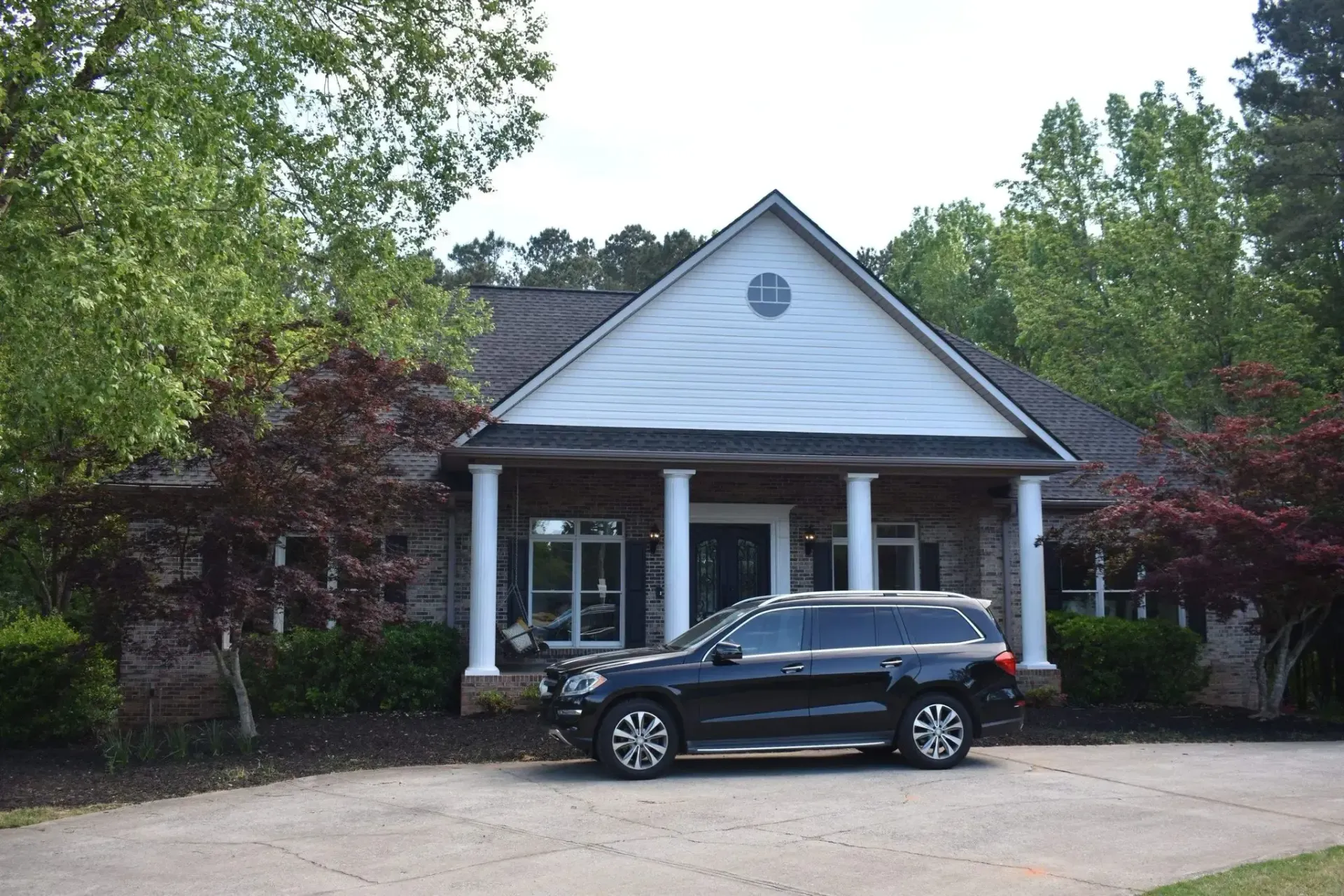 Black SUV parked in front of a brick house with white columns and a brown roof, trees in the background.