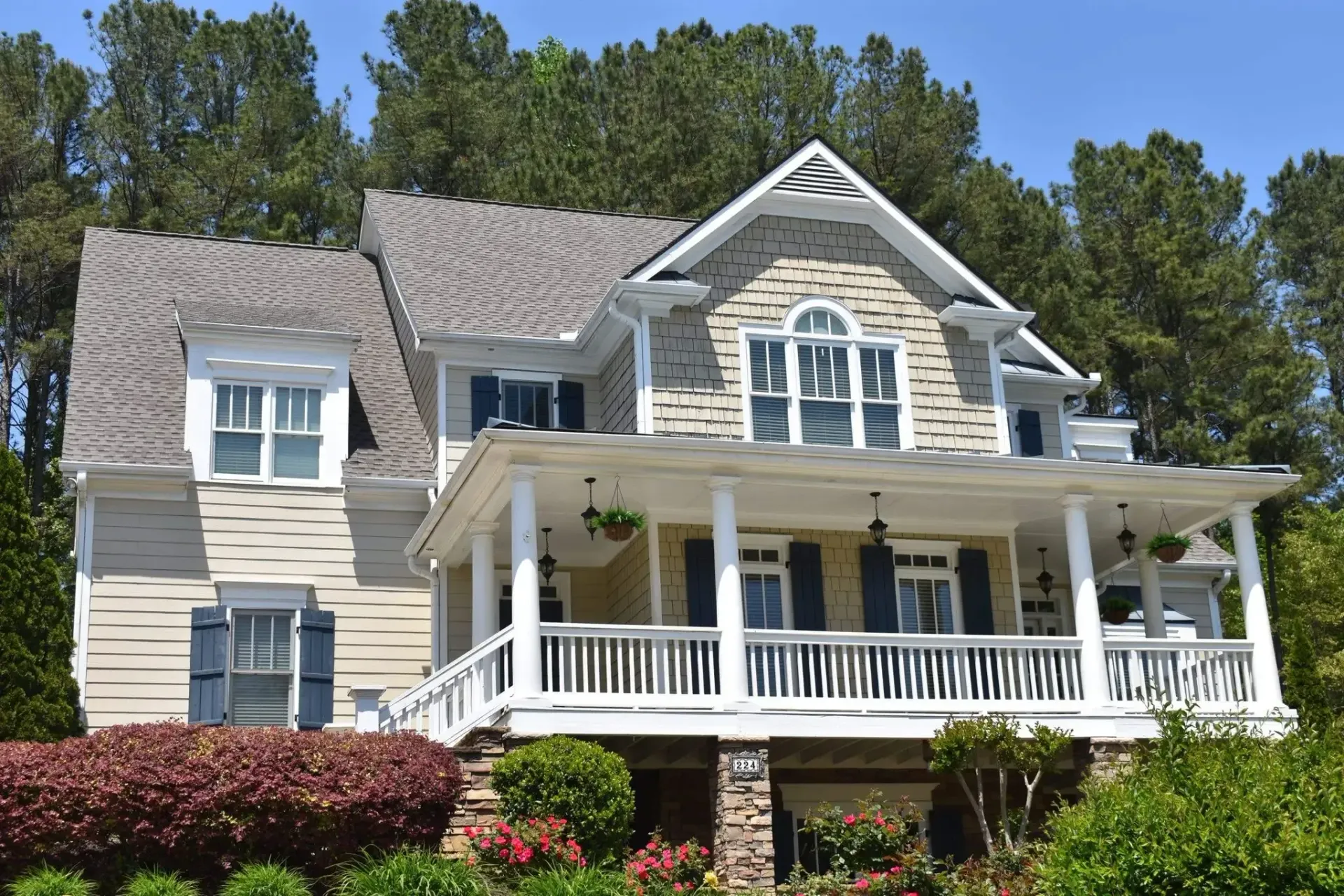 Two-story house with a wraparound porch, stone accents, and blue shutters, in front of trees.