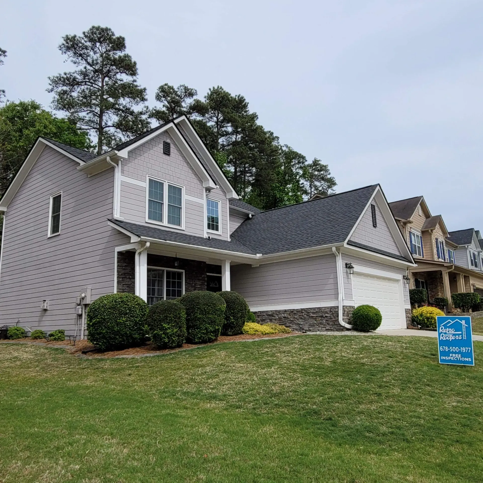 Gray house with white trim and bushes, another brick house visible on the right, and green grass.