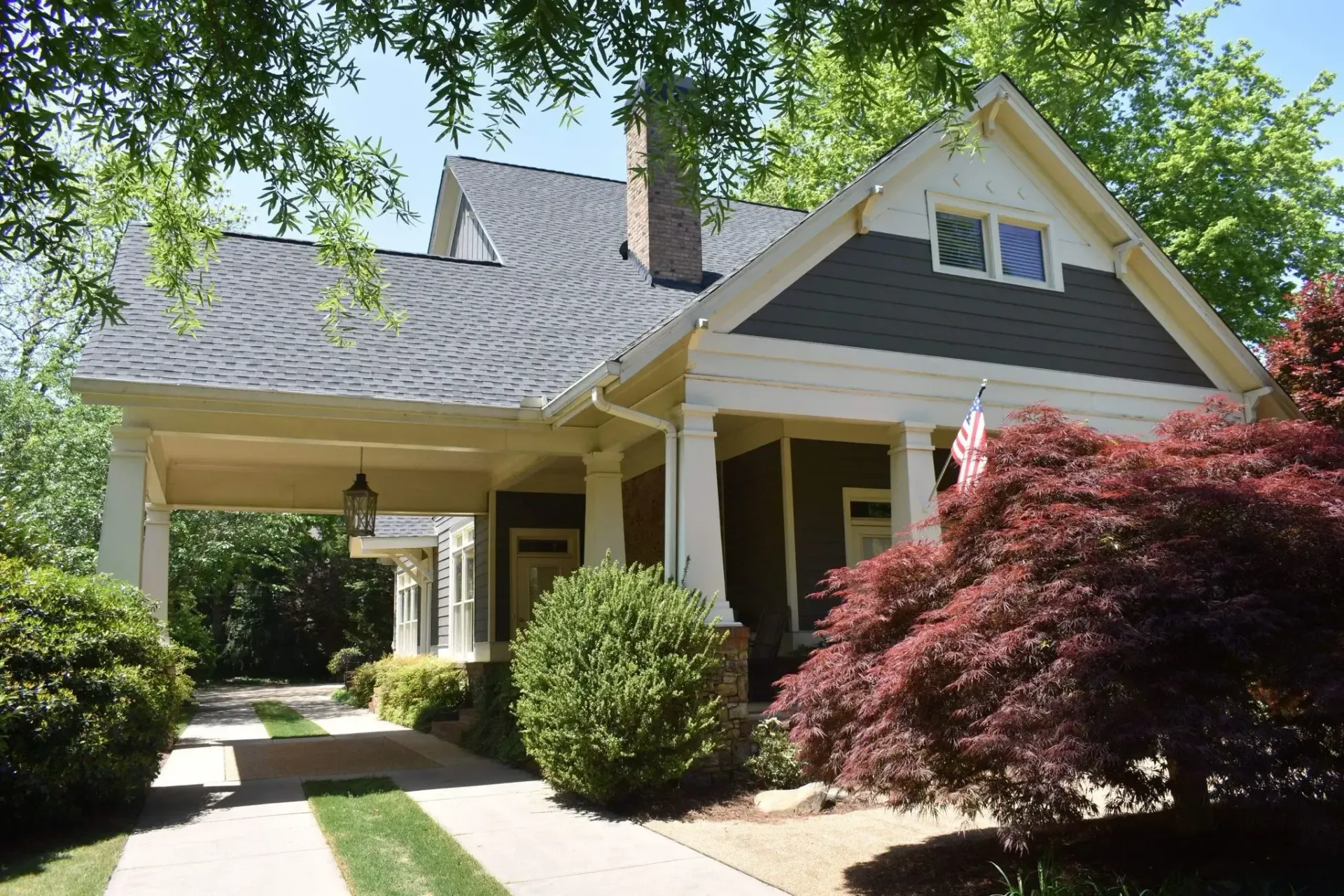 Craftsman-style house with a carport. Green lawn and bushes, red-leafed tree in front. Brick chimney.