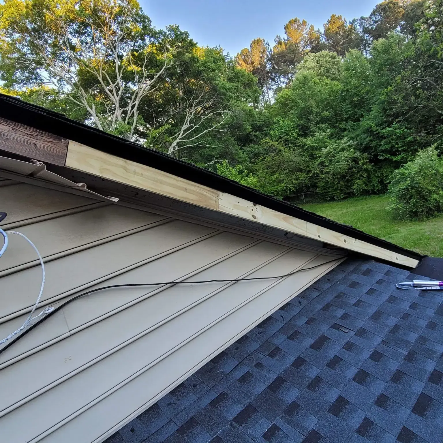 A roof edge with missing trim. Tan siding, blue shingles, and a forested background.