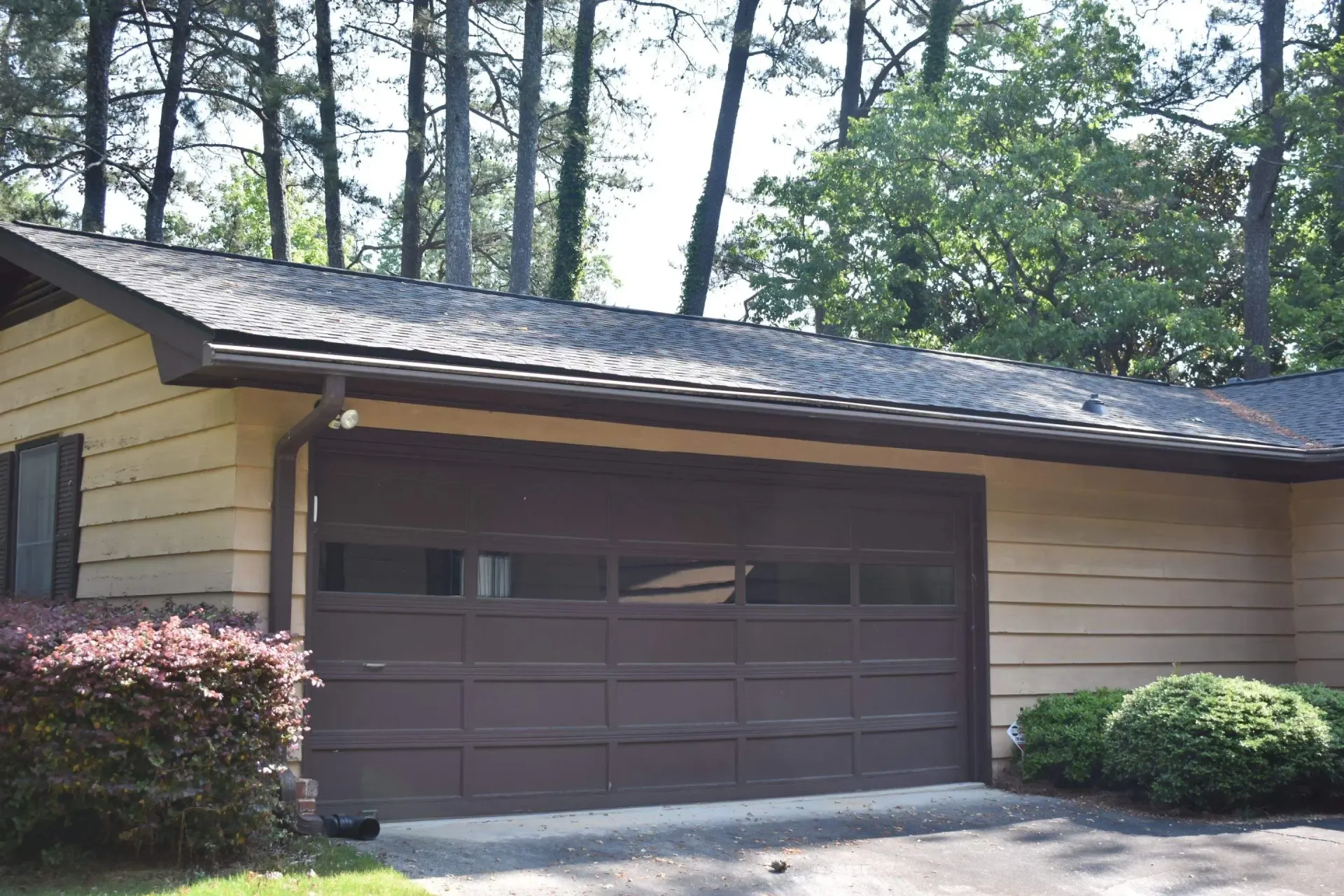Tan building with brown garage door, dark roof, bushes, trees.