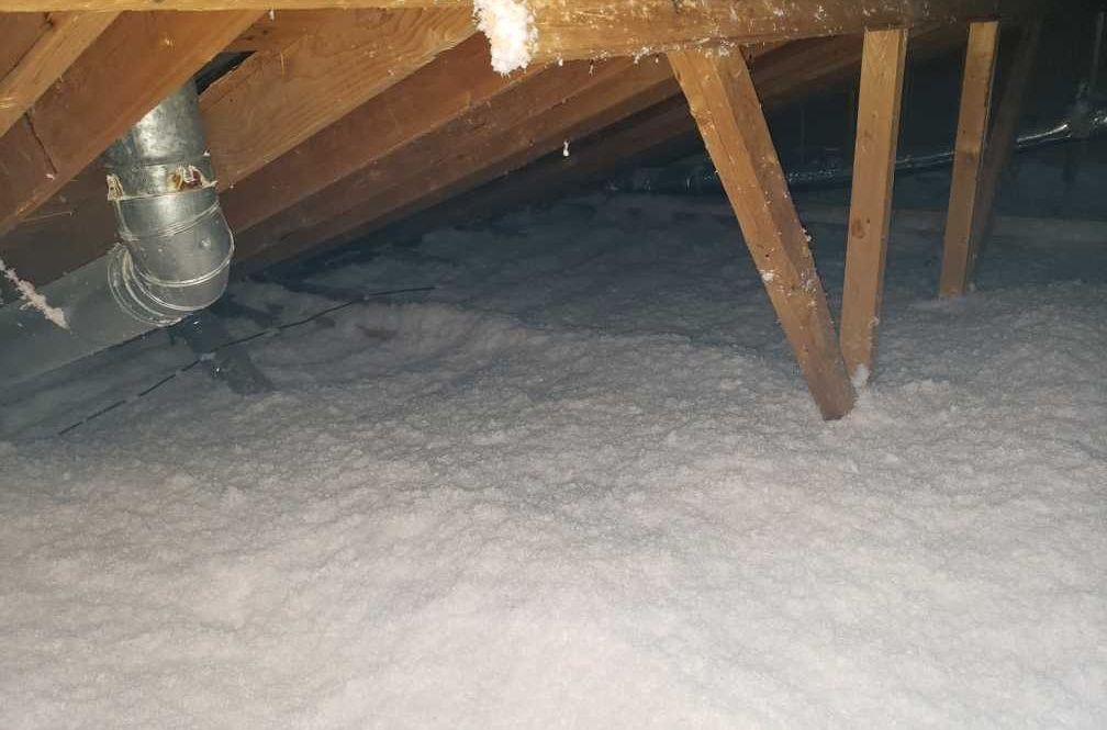 Attic with exposed wooden beams, insulation, and a duct, illuminated by overhead light.