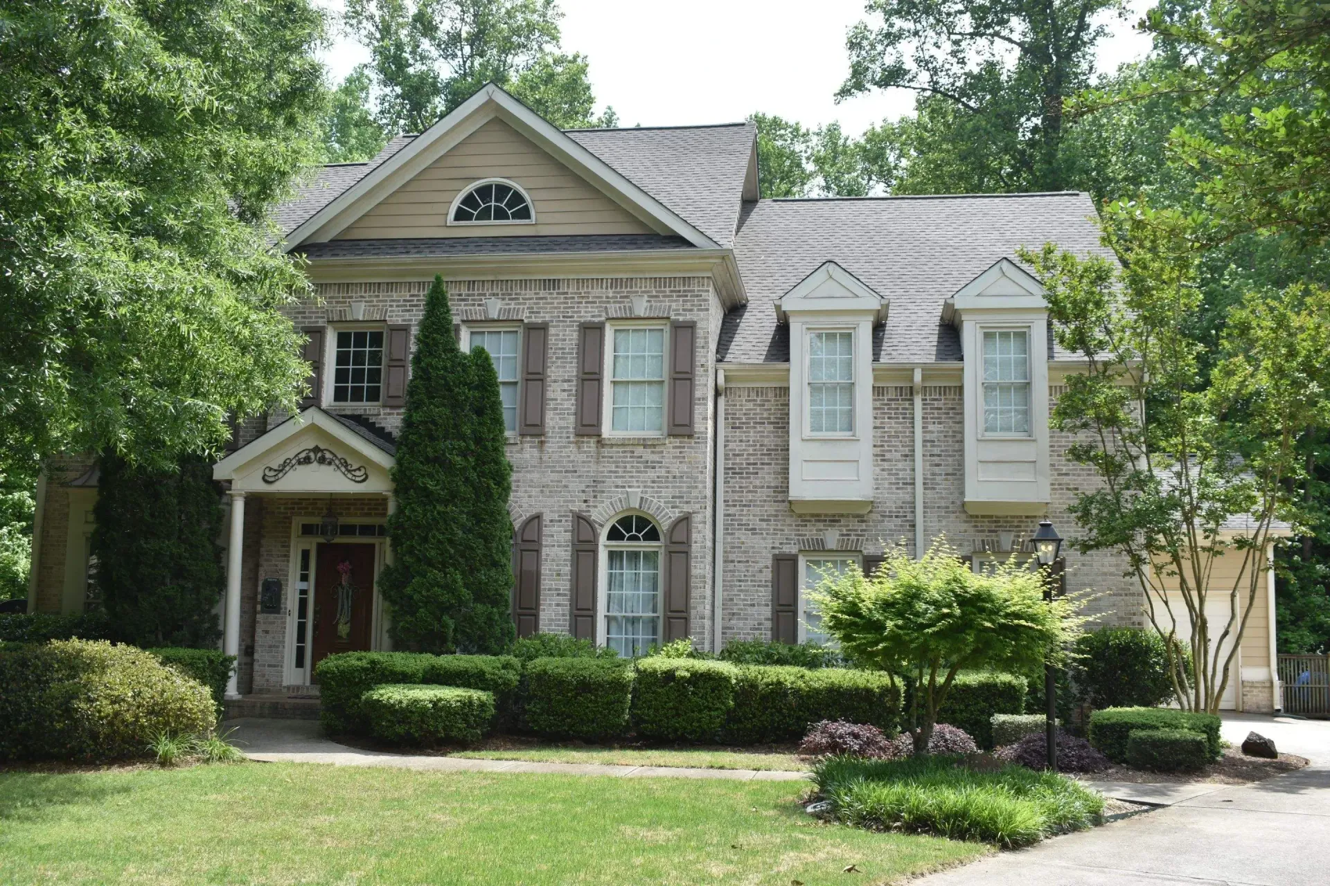 Two-story brick house with brown shutters, bay windows, and a well-manicured lawn, surrounded by trees.