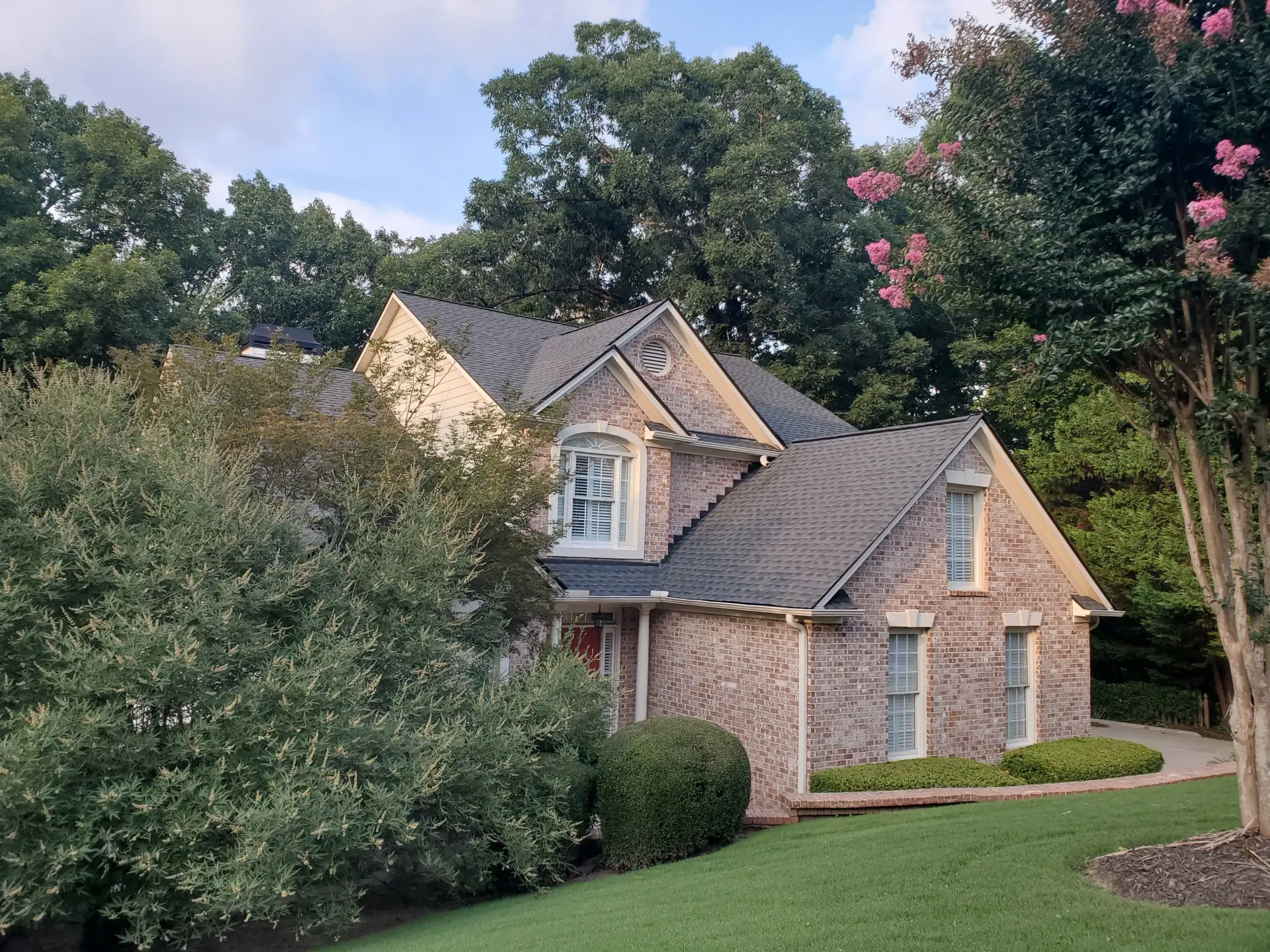 Brick house nestled in greenery; light-colored brick, dark roof, and arched window, partially covered by foliage.