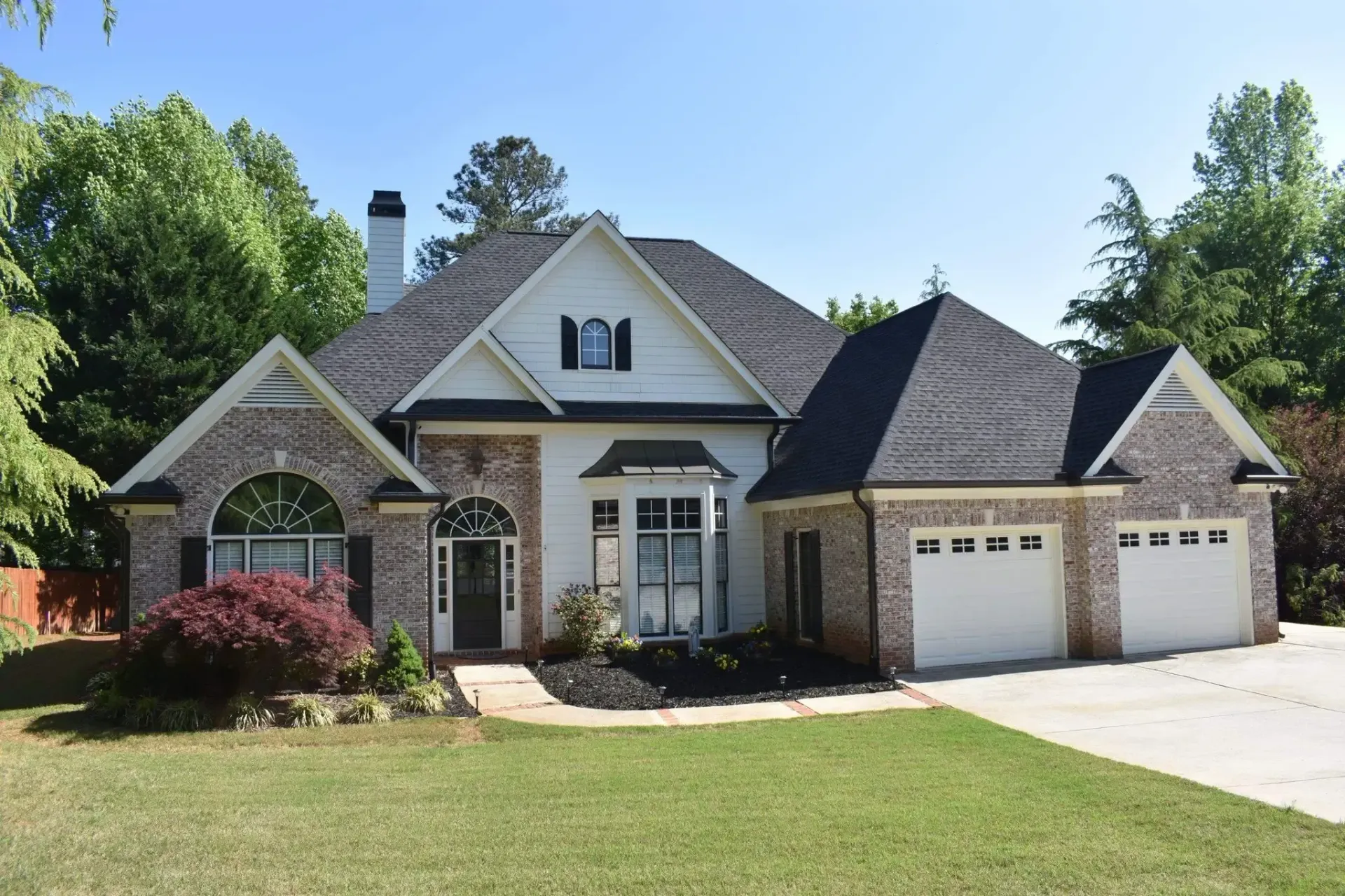 White house with brick accents, black roof, and three-car garage, set on a green lawn.
