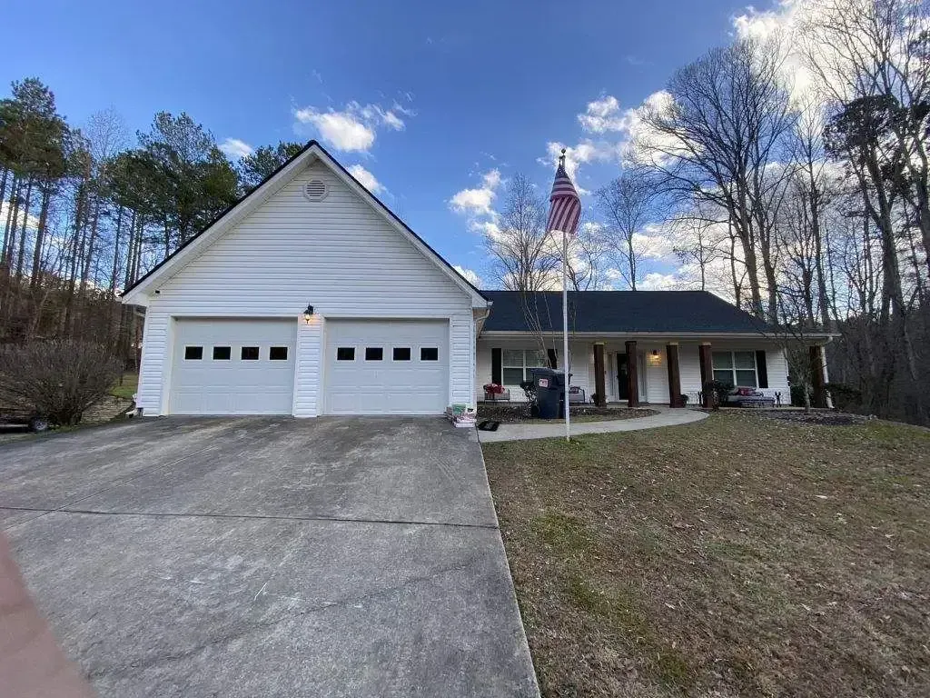 White house with a two-car garage, American flag, and a driveway. Blue sky, trees.