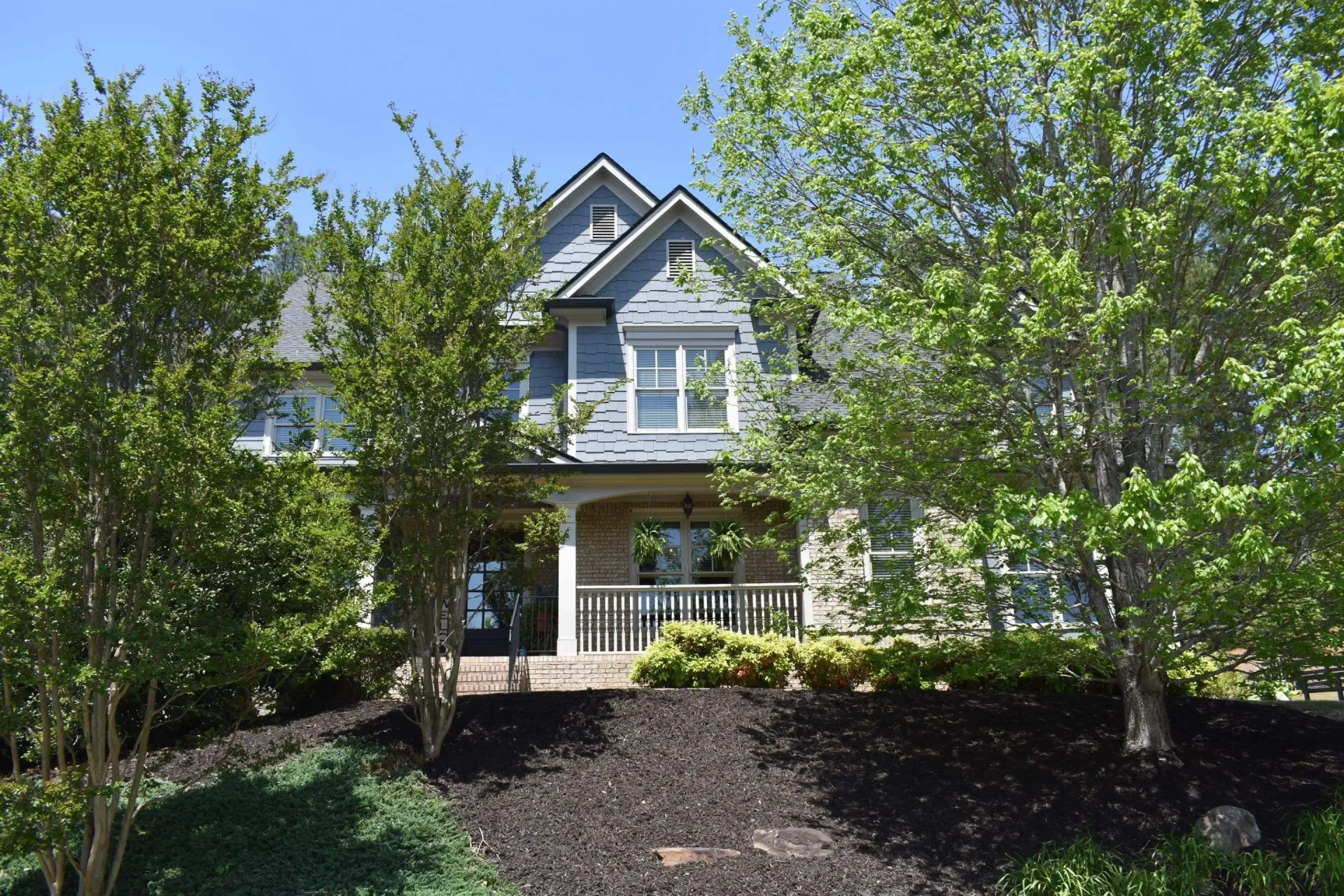 Two-story house with blue siding and a porch, partially obscured by green trees, on a sunny day.