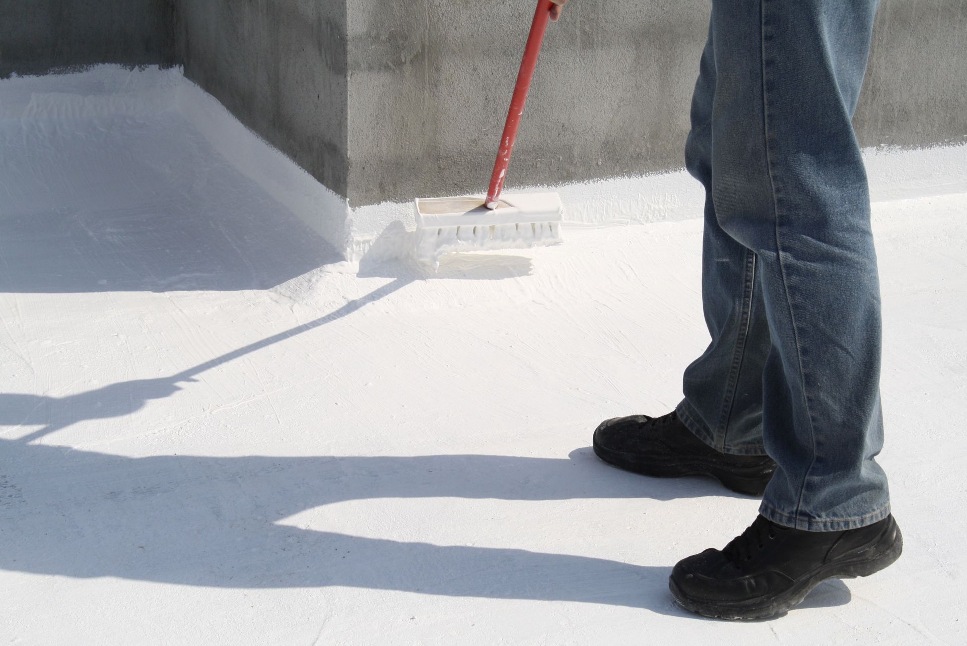 Person applying white coating to a flat roof with a long-handled brush.