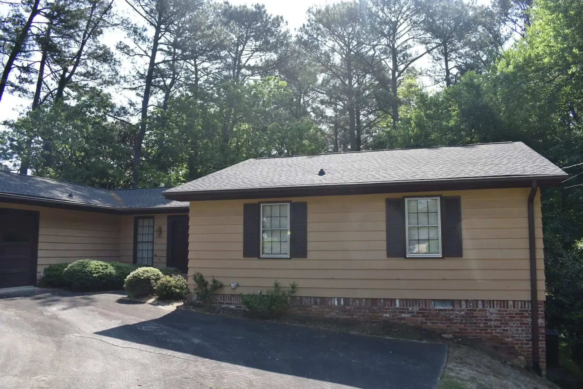 Beige house with dark shutters and roof, set in front of trees.