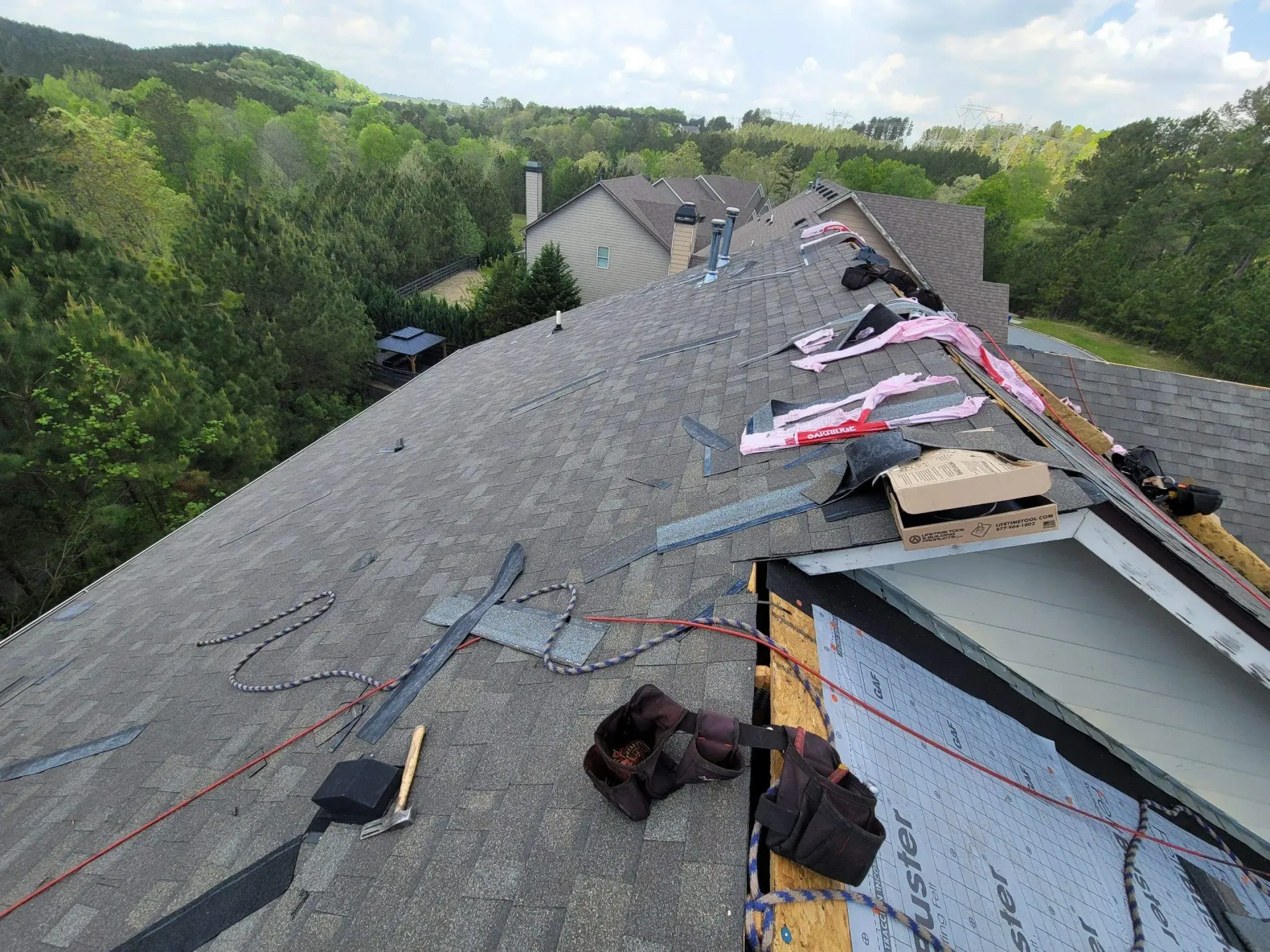 Roof of a house under construction; shingles removed, tools and insulation visible. Trees in background.