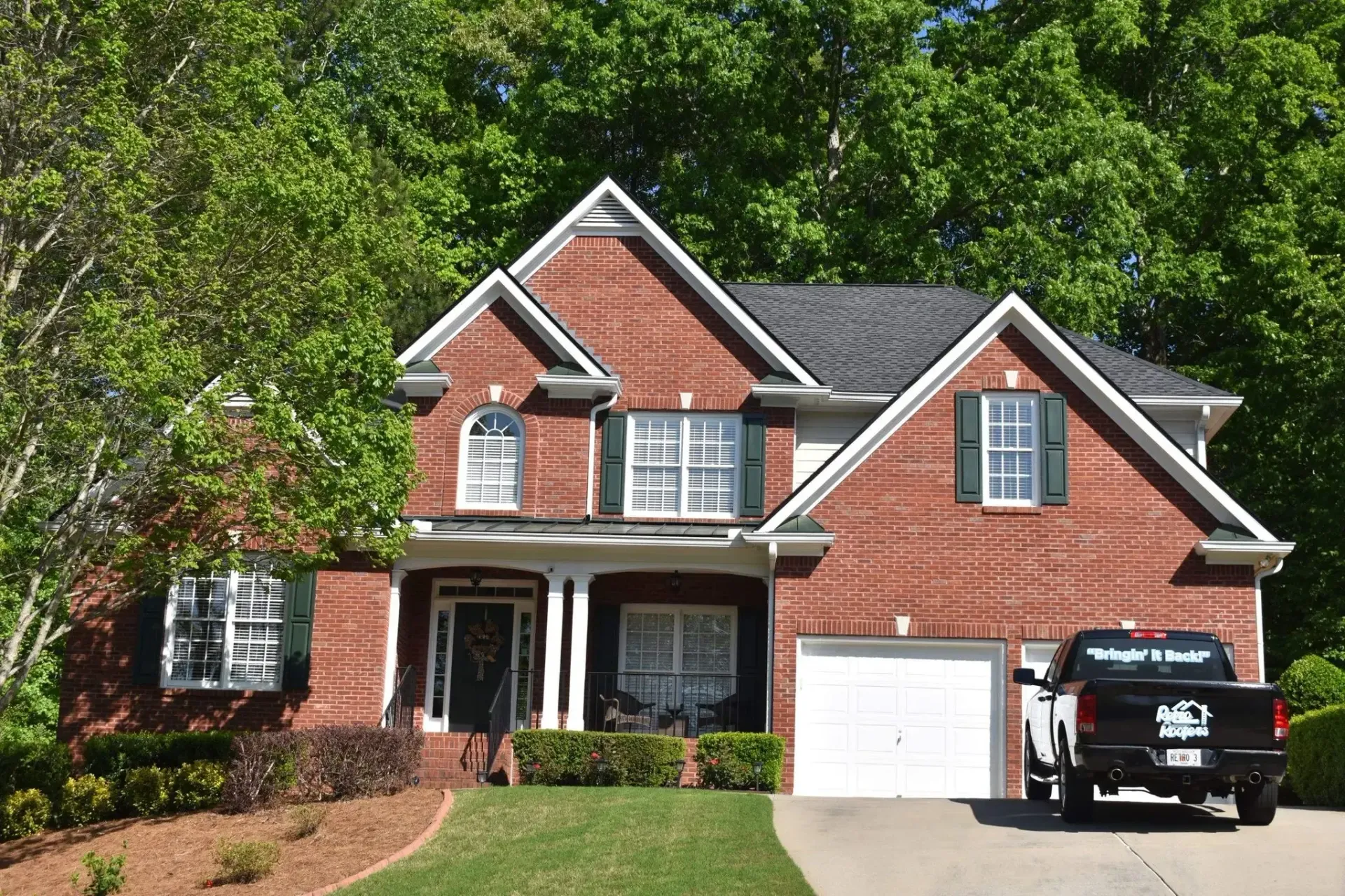 Red brick house with white trim, green shutters, and a black truck in the driveway, in a tree-lined setting.