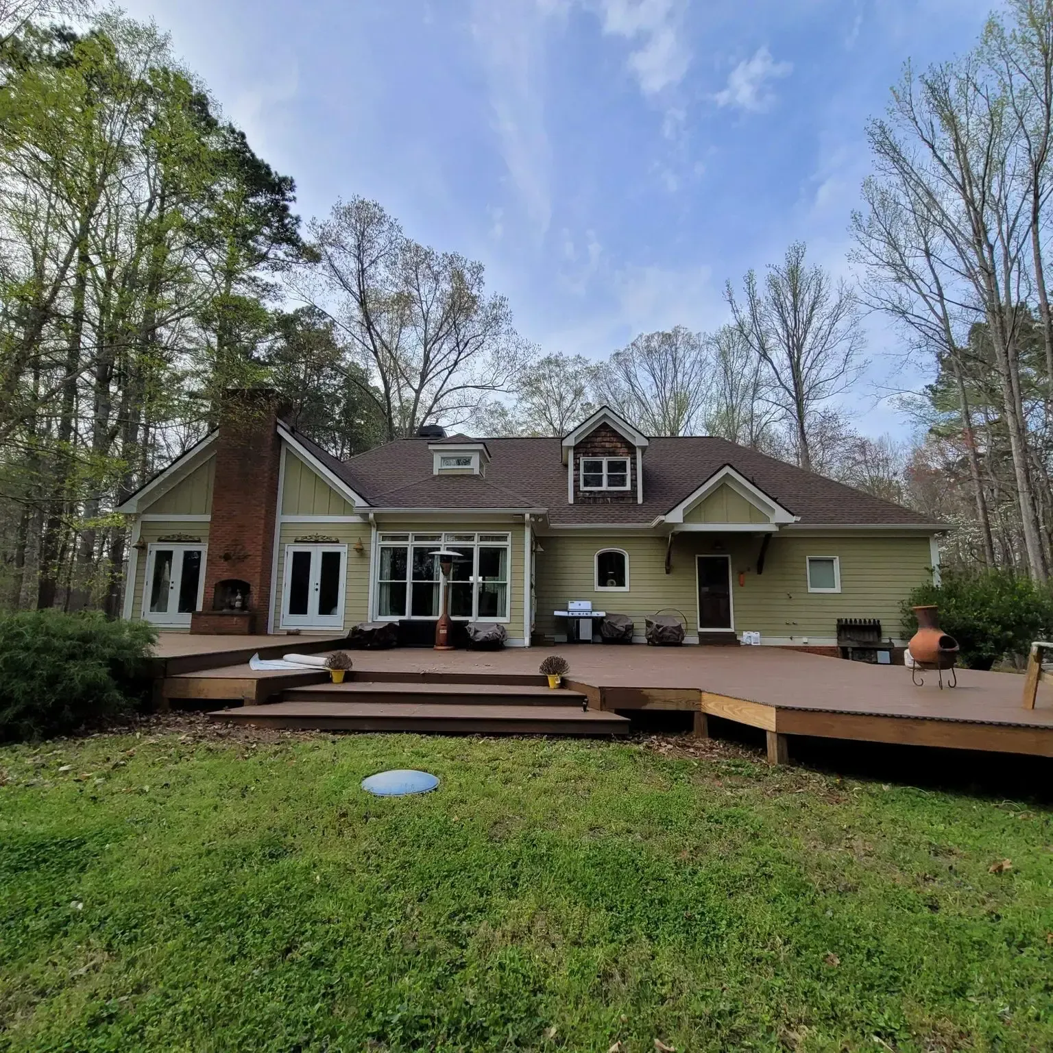A light yellow house with a brown roof and large wooden deck sits in a grassy yard surrounded by trees.
