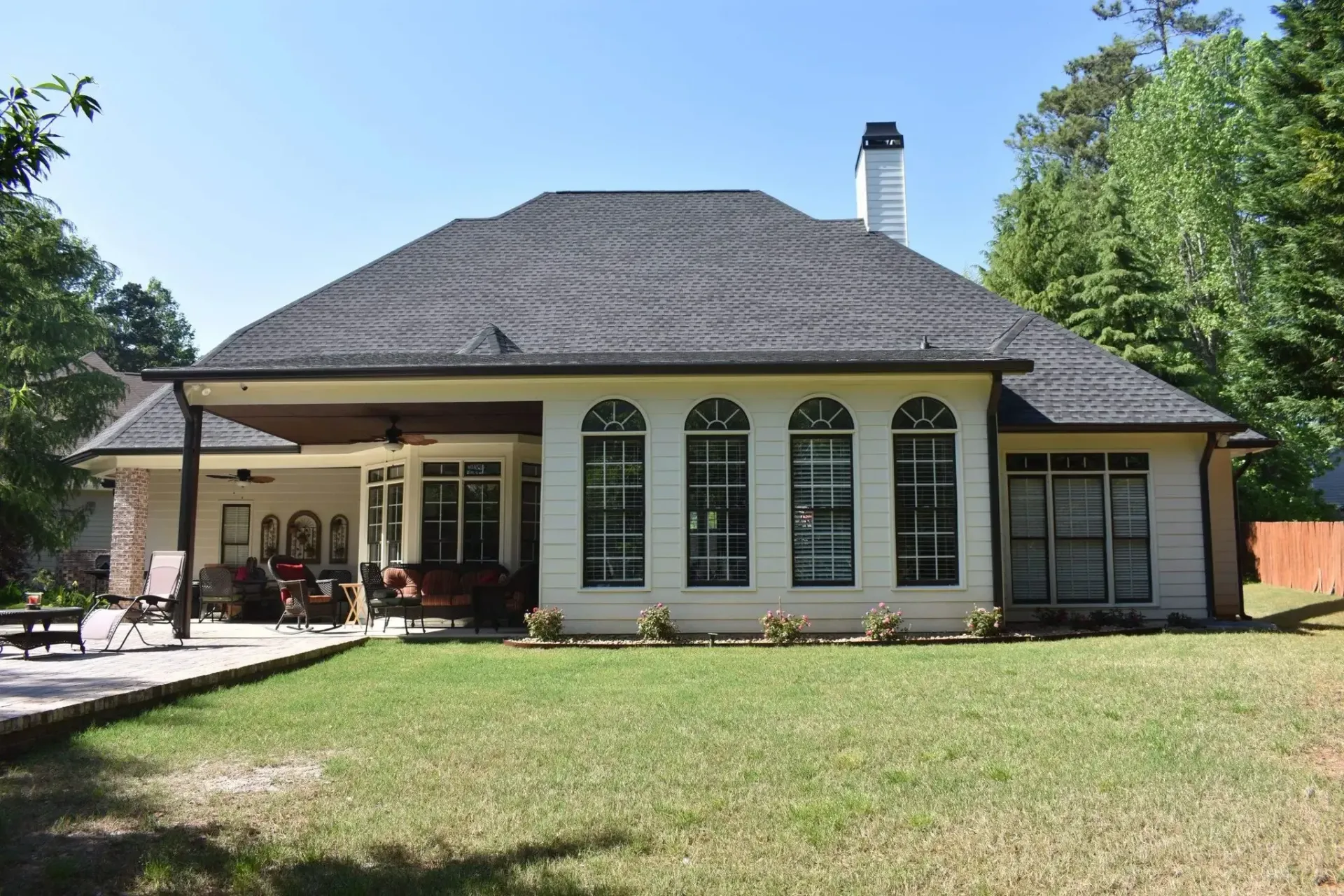 Backyard view of a beige house with black roof, arched windows, and covered patio.
