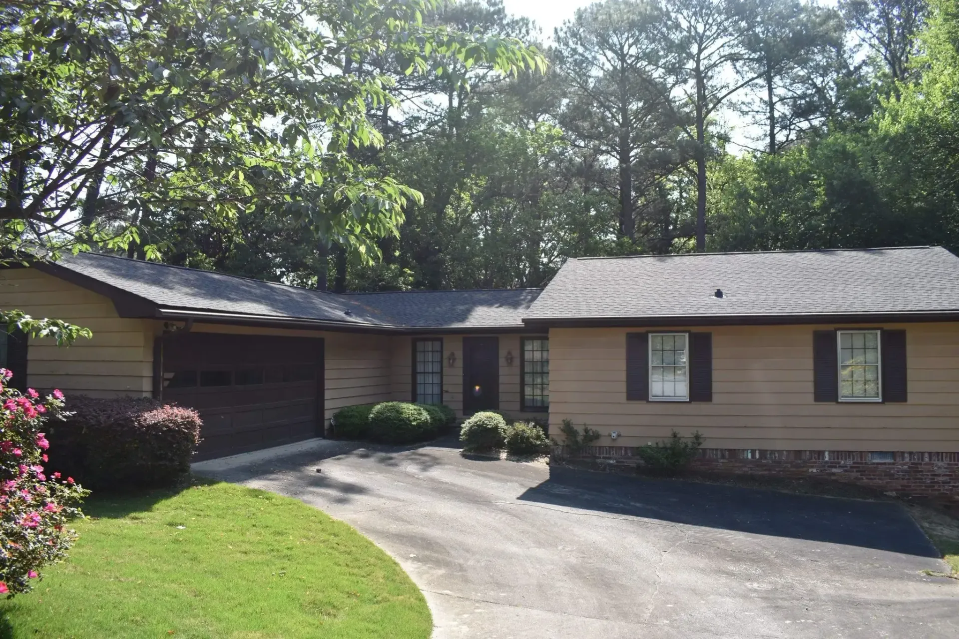 Beige ranch-style house with a dark roof, driveway, and greenery, with a garage and shutters.
