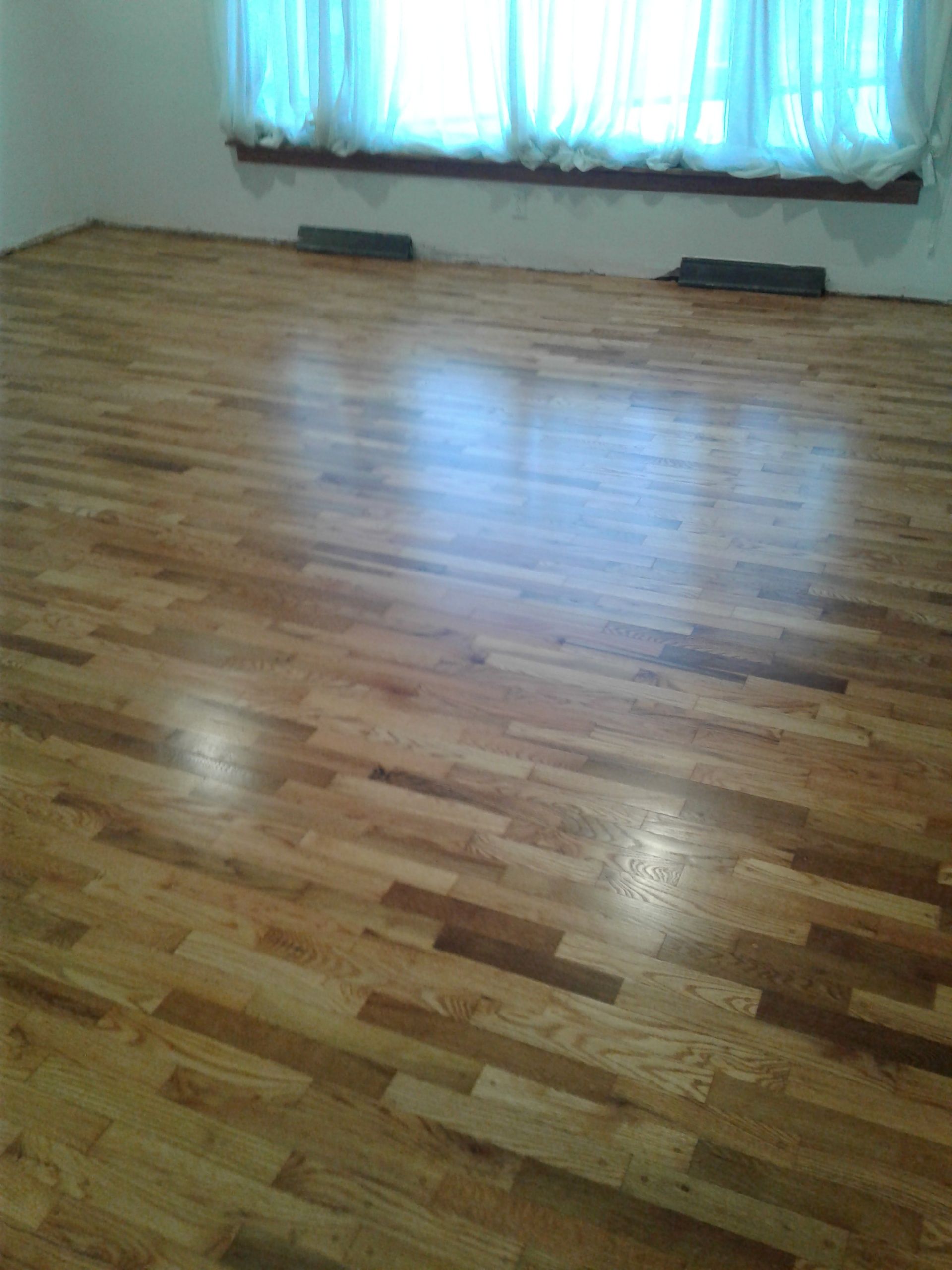 Hardwood floors with a mix of light and dark brown planks in a room with white walls and sheer curtains.