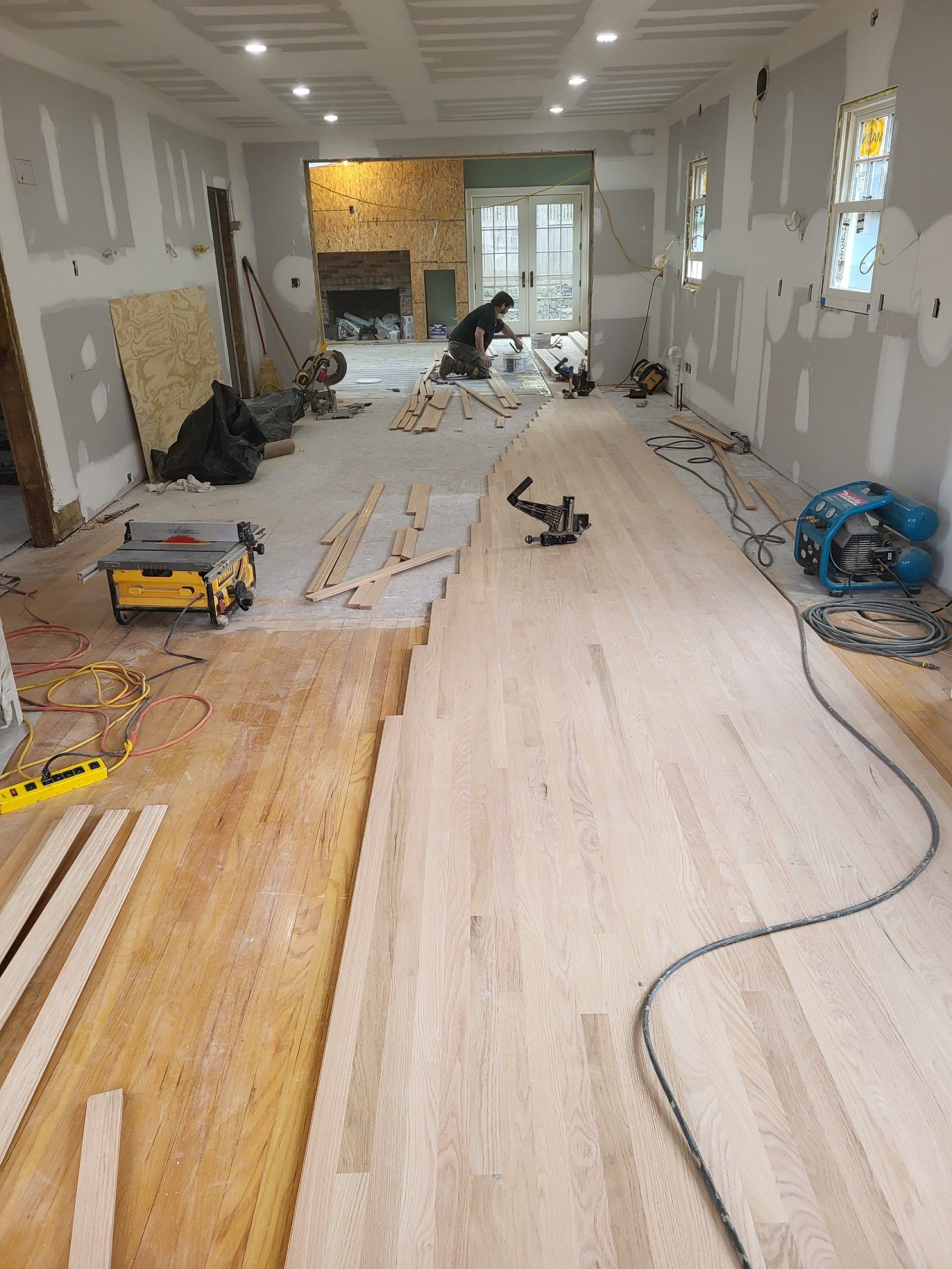 A person installs light-colored hardwood flooring in a room under construction with bare walls and tools scattered around.