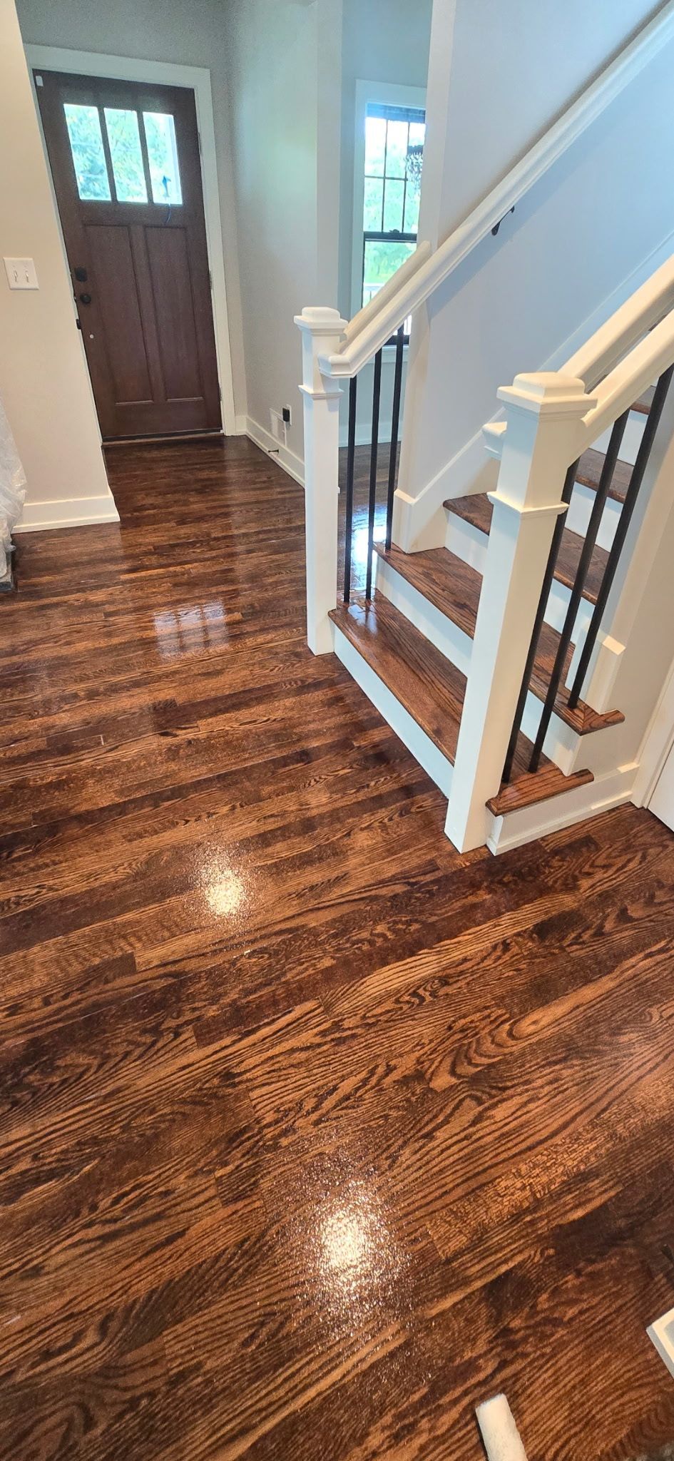 Polished hardwood floors in a hallway leading to a dark wooden front door and a white staircase with a metal railing.