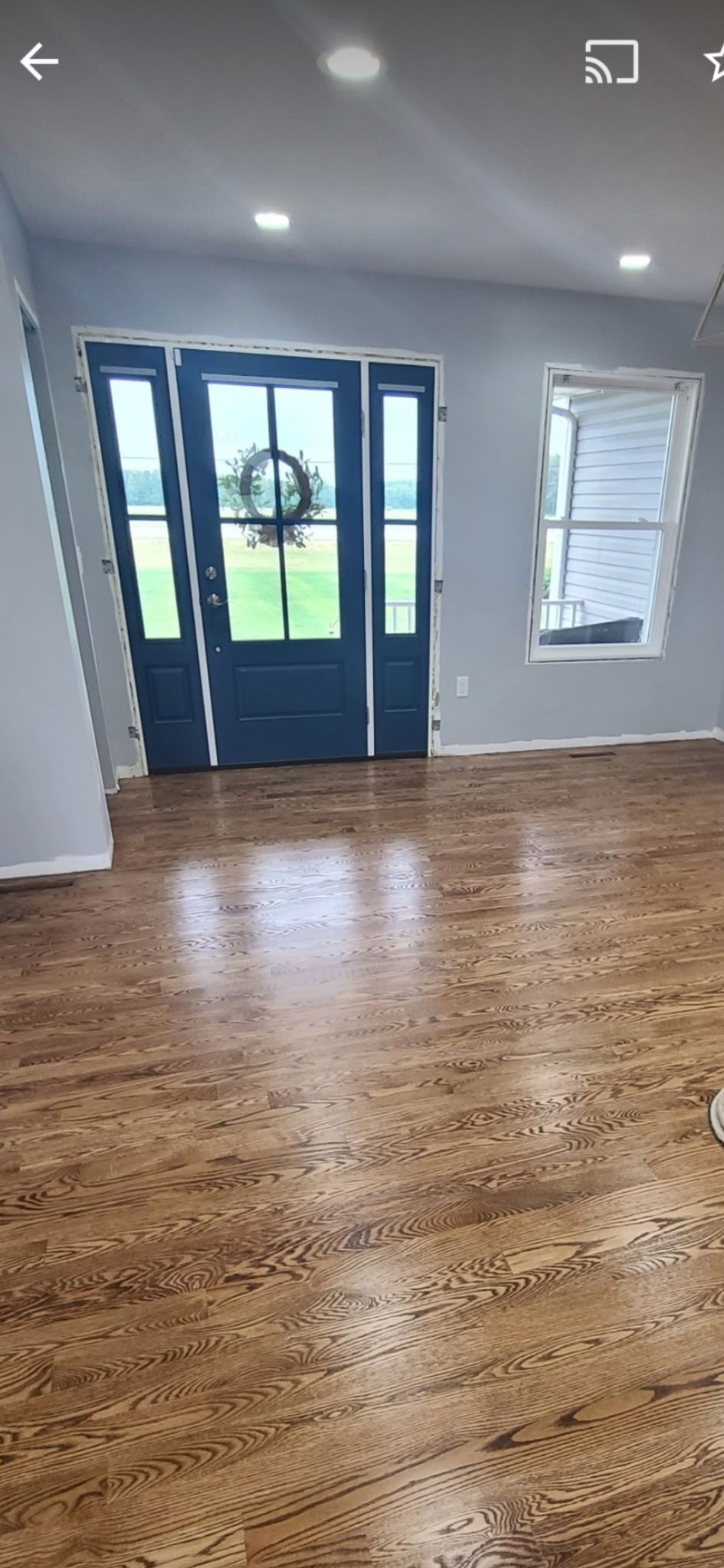 A light grey room with polished hardwood floors, a dark blue front door with side lights, and a window with white blinds.