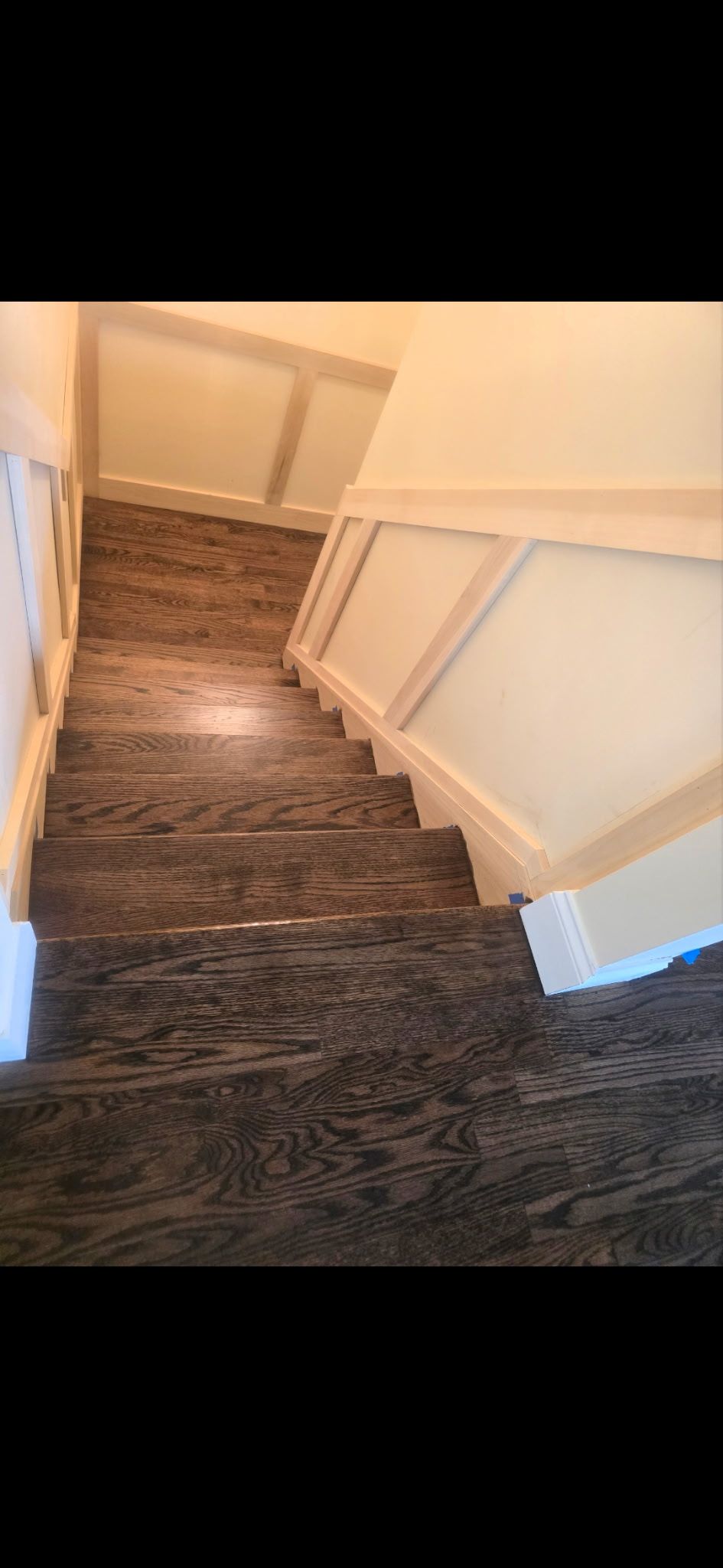 A downward view of a wooden staircase with dark, grain-patterned steps flanked by off-white wainscoting and trim.