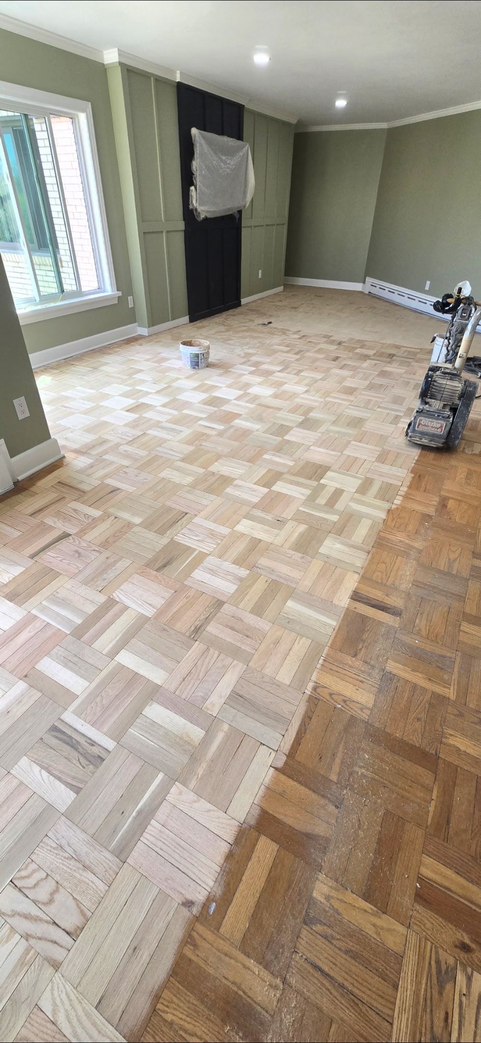 A room with parquet flooring, half of which is sanded down to raw wood, with a sanding machine nearby.