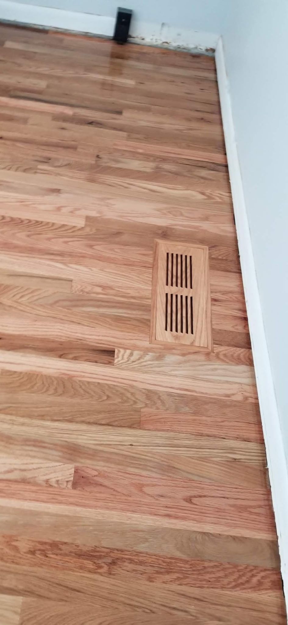 A light-colored hardwood floor featuring a rectangular wooden air vent, situated next to a white wall baseboard.
