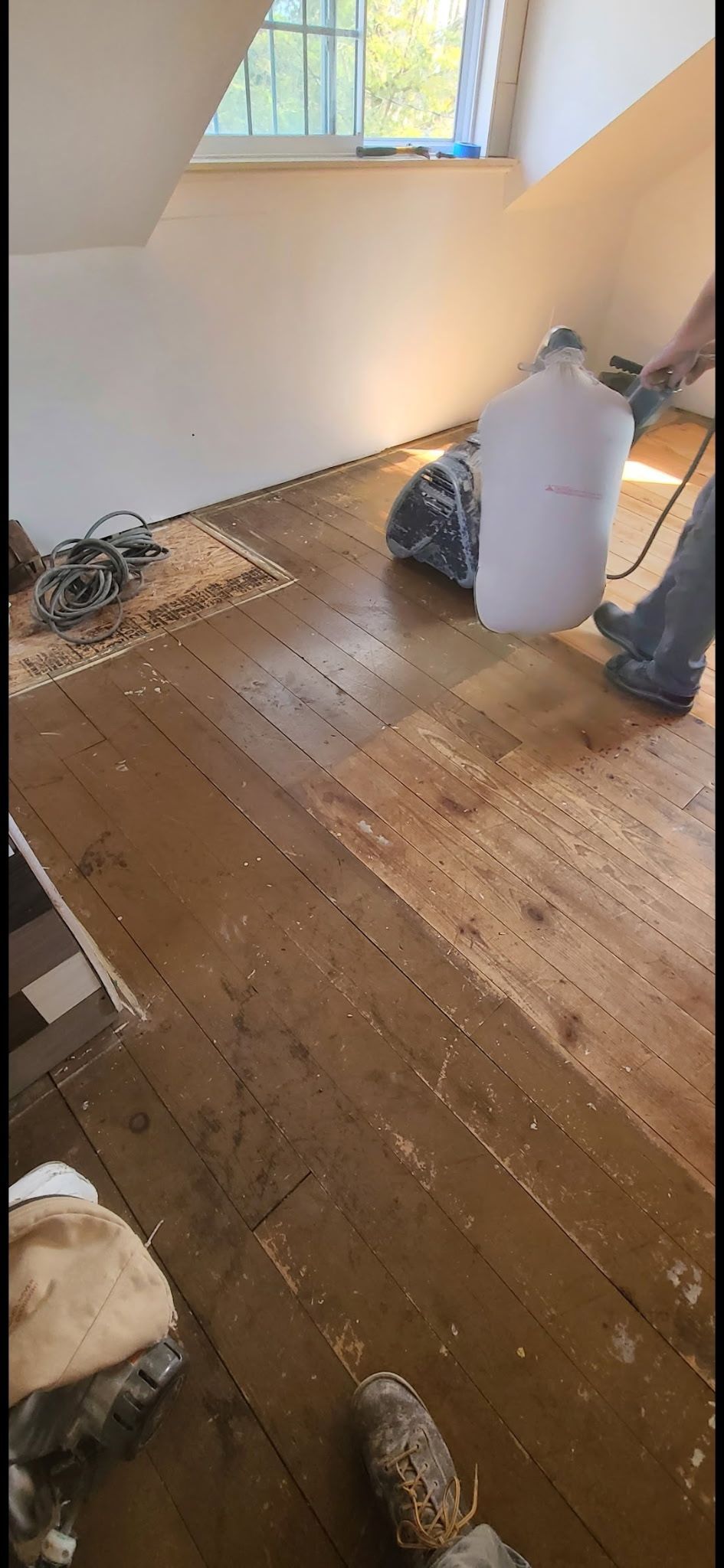 A person uses a floor sander on old wooden floorboards in a room with a sloped ceiling and window.