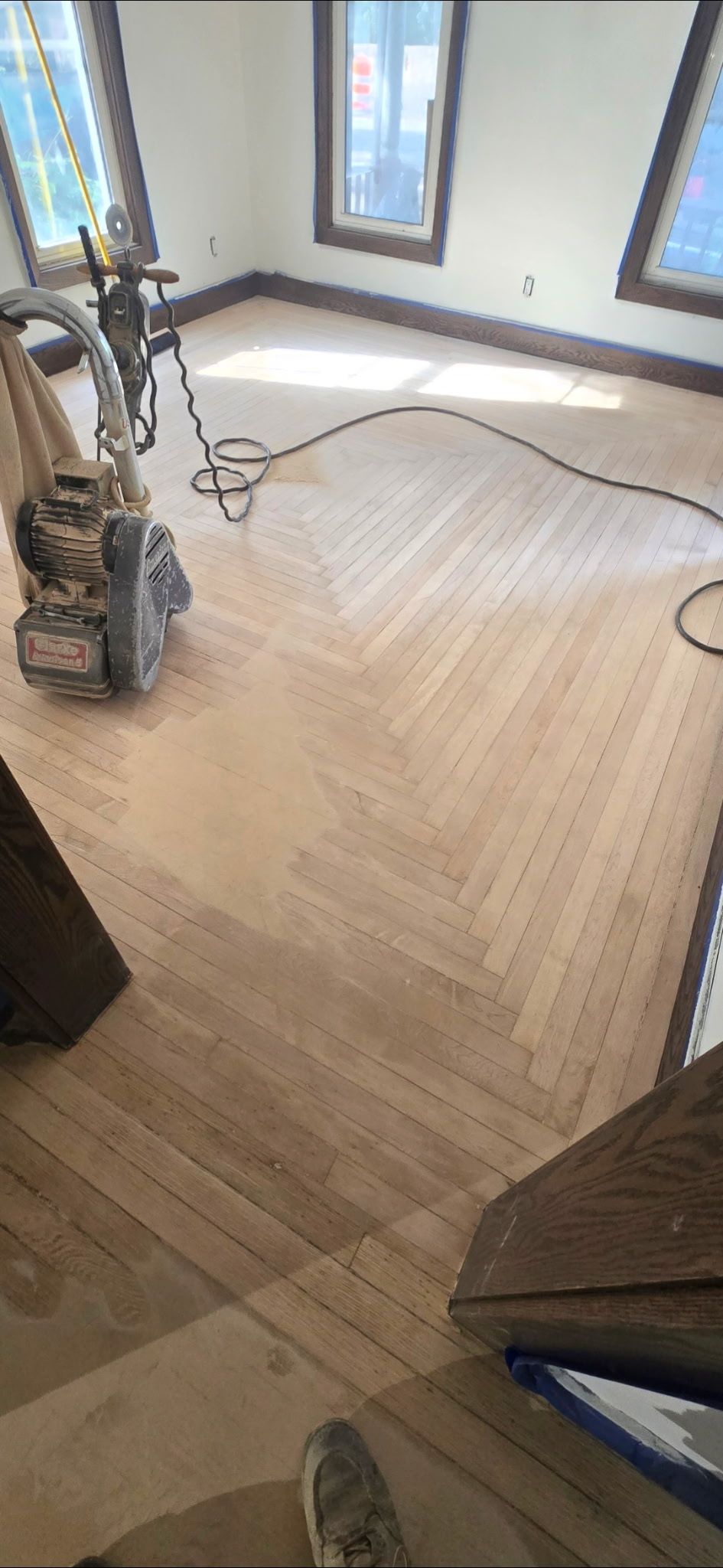 A floor sanding machine sits on a partially sanded herringbone hardwood floor in a room with three large windows.