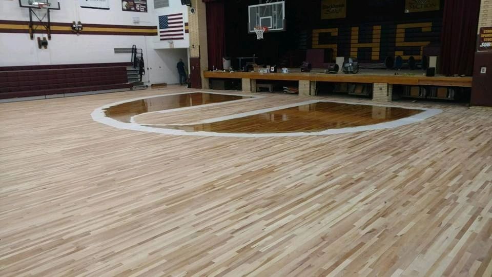 A basketball court with light-colored, sanded wood flooring and a darker, polished semi-circle area near the stage.