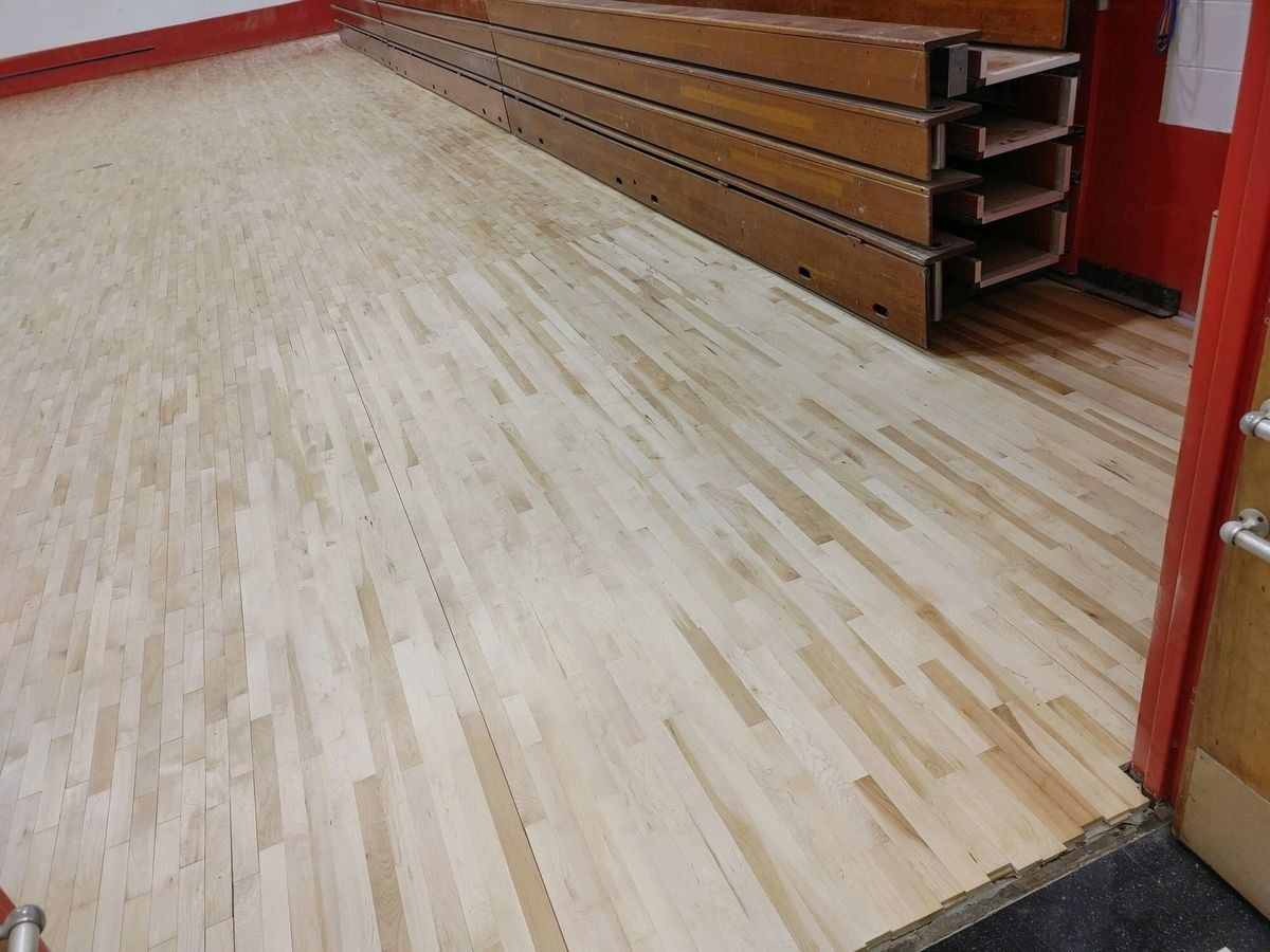 A light-colored wood floor with a diagonal pattern next to folded wooden bleachers in a gym or school hall.