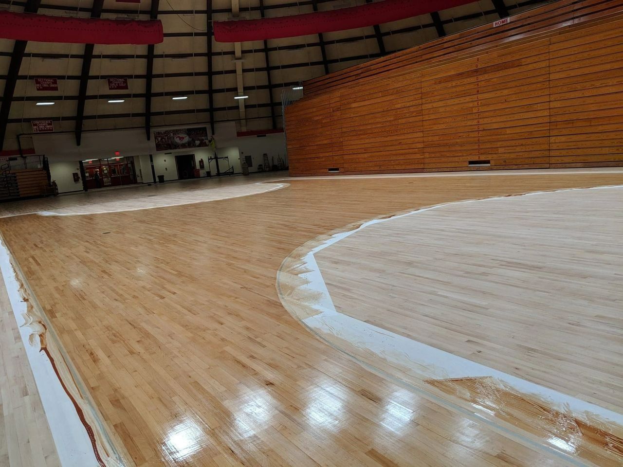 Indoor sports arena with a wooden basketball floor partially finished with a glossy sealant.