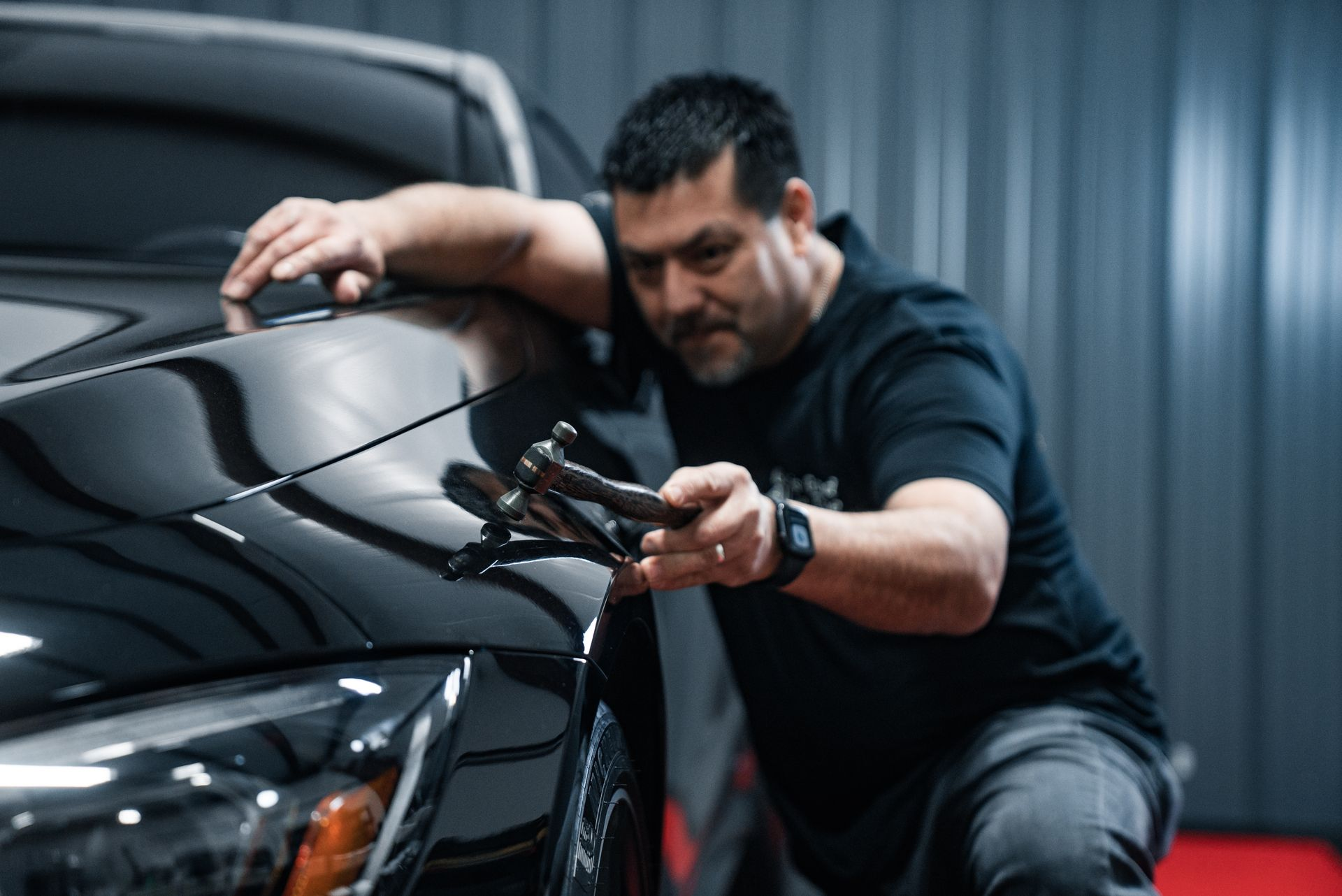 Raul Lara using blending hammer on a black car's front fender in a garage.