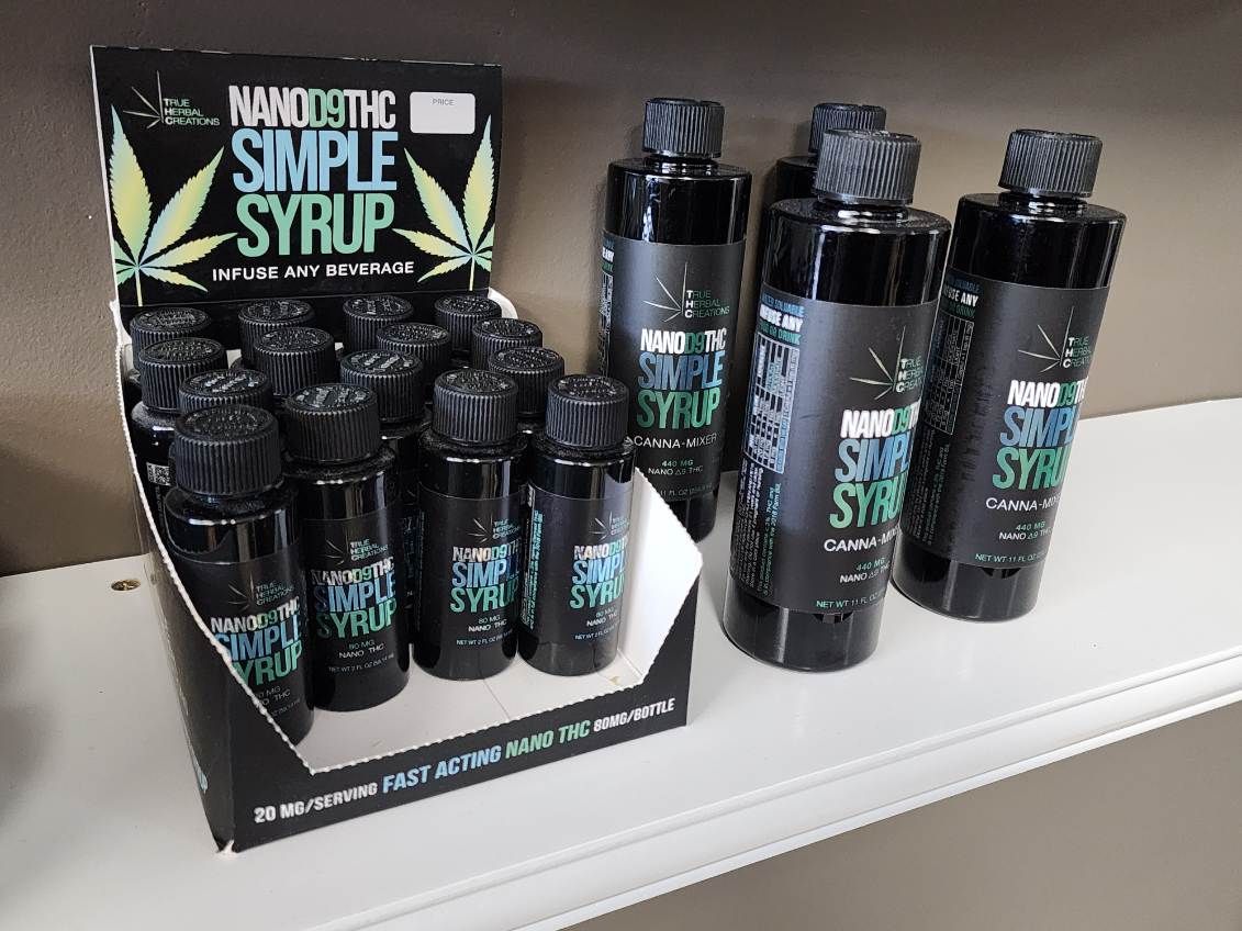 Display of NanoDose Simple Syrup bottles in a store setting. The bottles are black with a turquoise and white label, in a cardboard display.