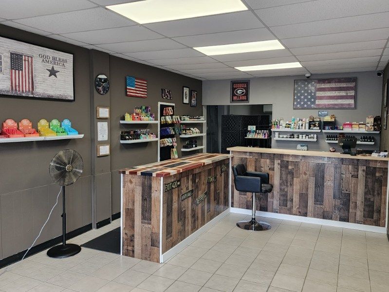 Interior of a vape shop with wood-paneled counters, displays of products, and American flag decor. A stand-up fan is in the corner.