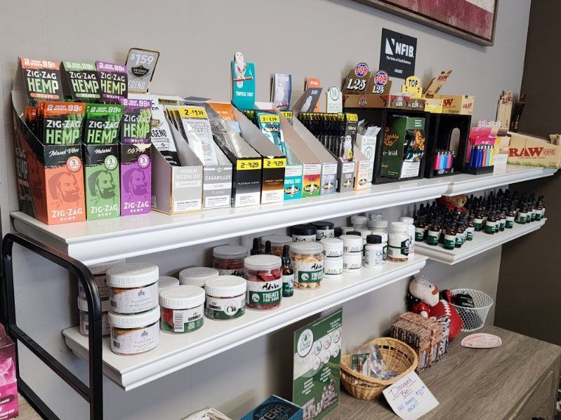 Shelves stocked with various cannabis products in a retail store. Products include jars, boxes, and pens of different brands.