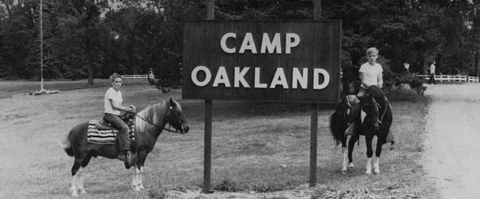 Two people are riding horses in front of a sign for camp oakland – Oxford, MI - Camp Oakland