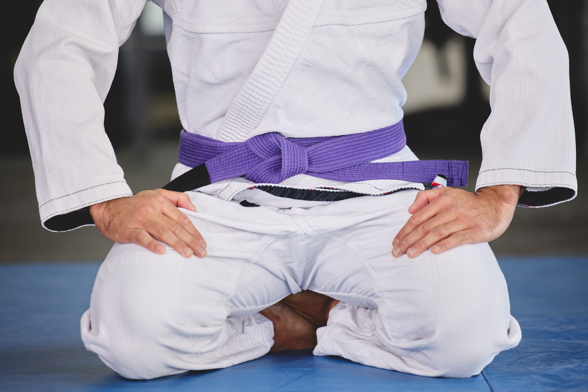A person in a white karate uniform with a purple belt is kneeling on a blue mat.