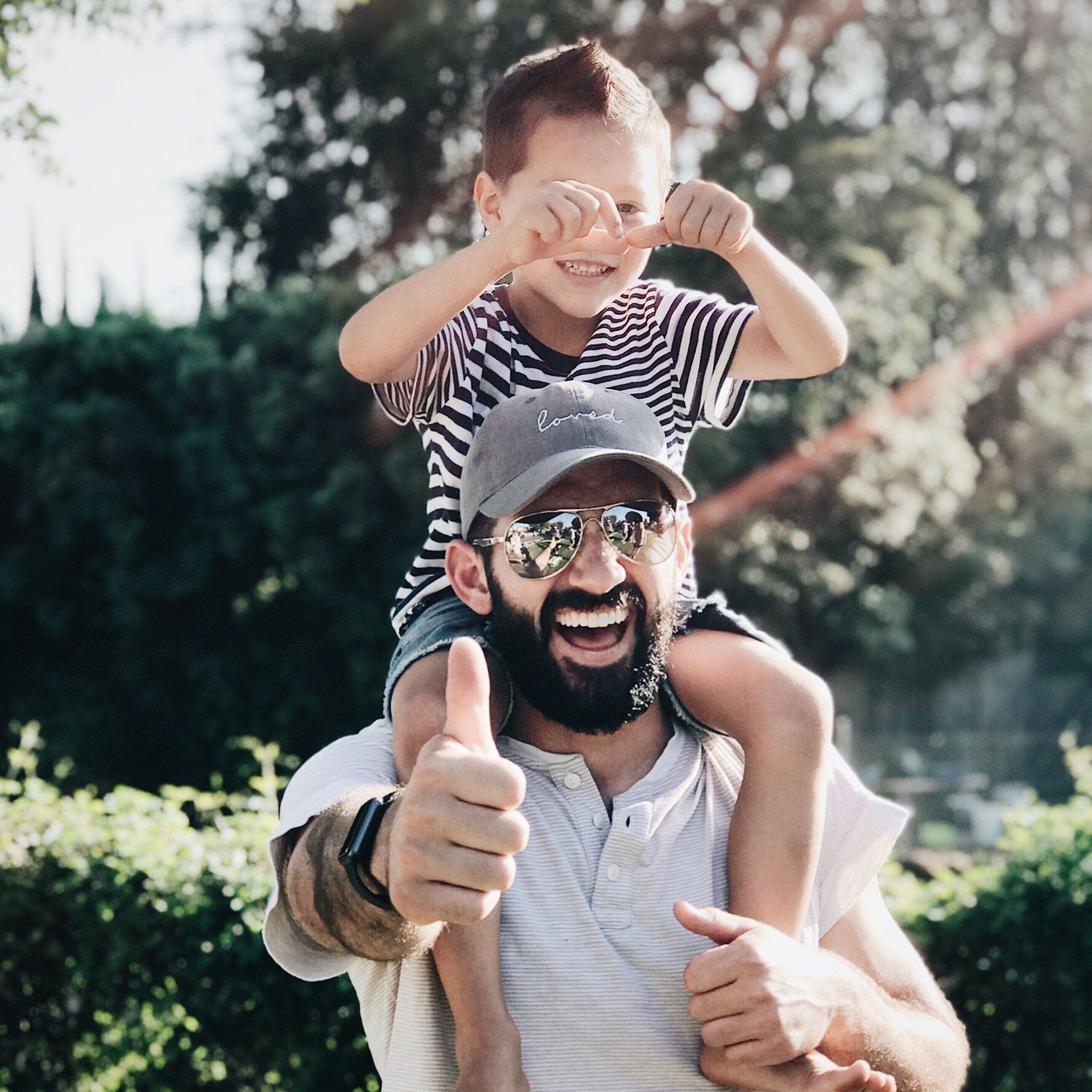 A man is carrying a little boy on his shoulders and giving a thumbs up.