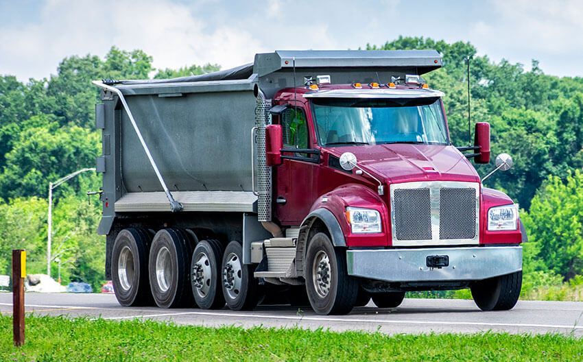 A red dump truck is driving down a road next to a grassy field.