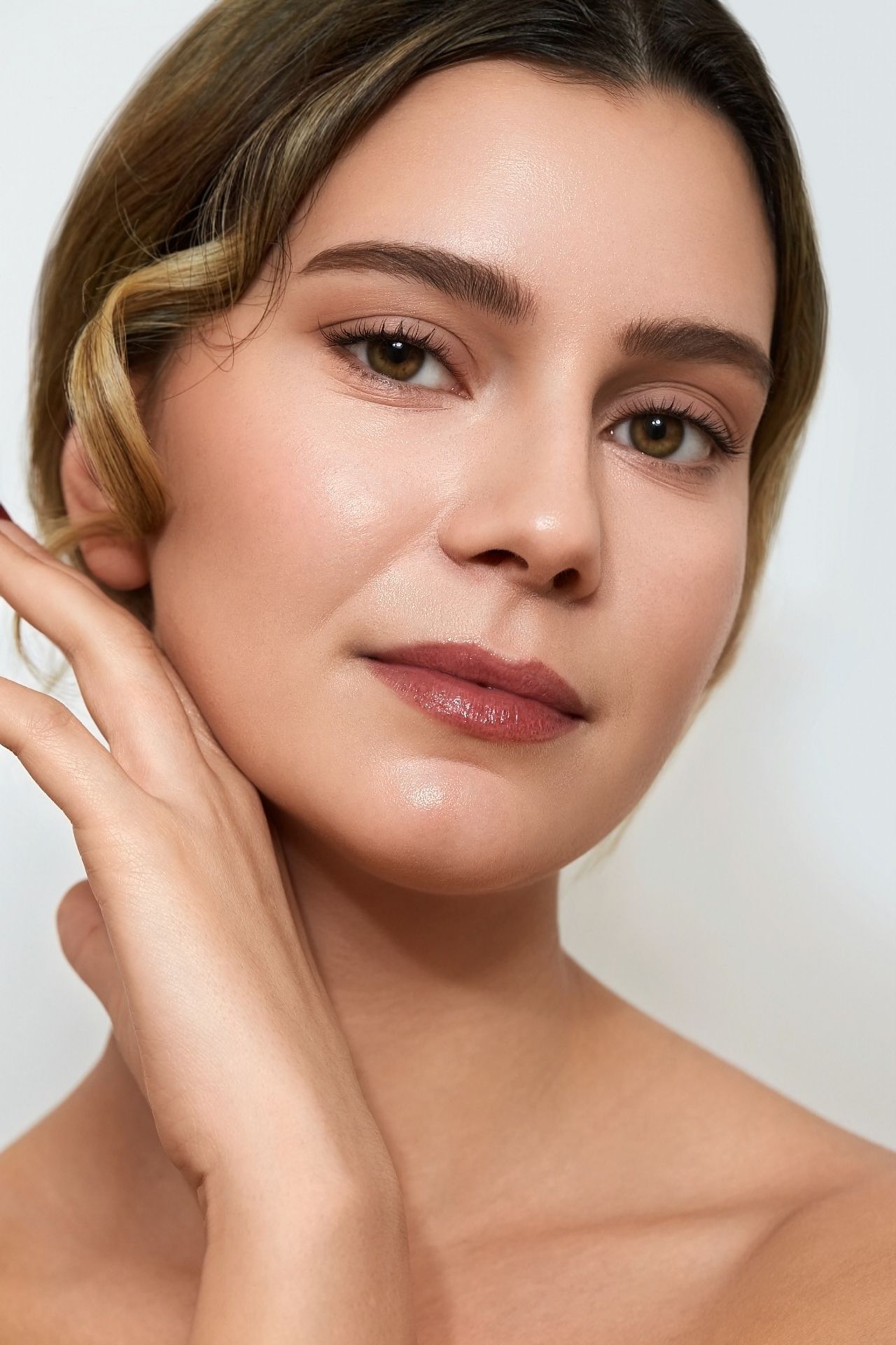 Frau mit heller Haut, hellbraunem Haar, geschminkt, berührt ihr Gesicht mit der Hand.