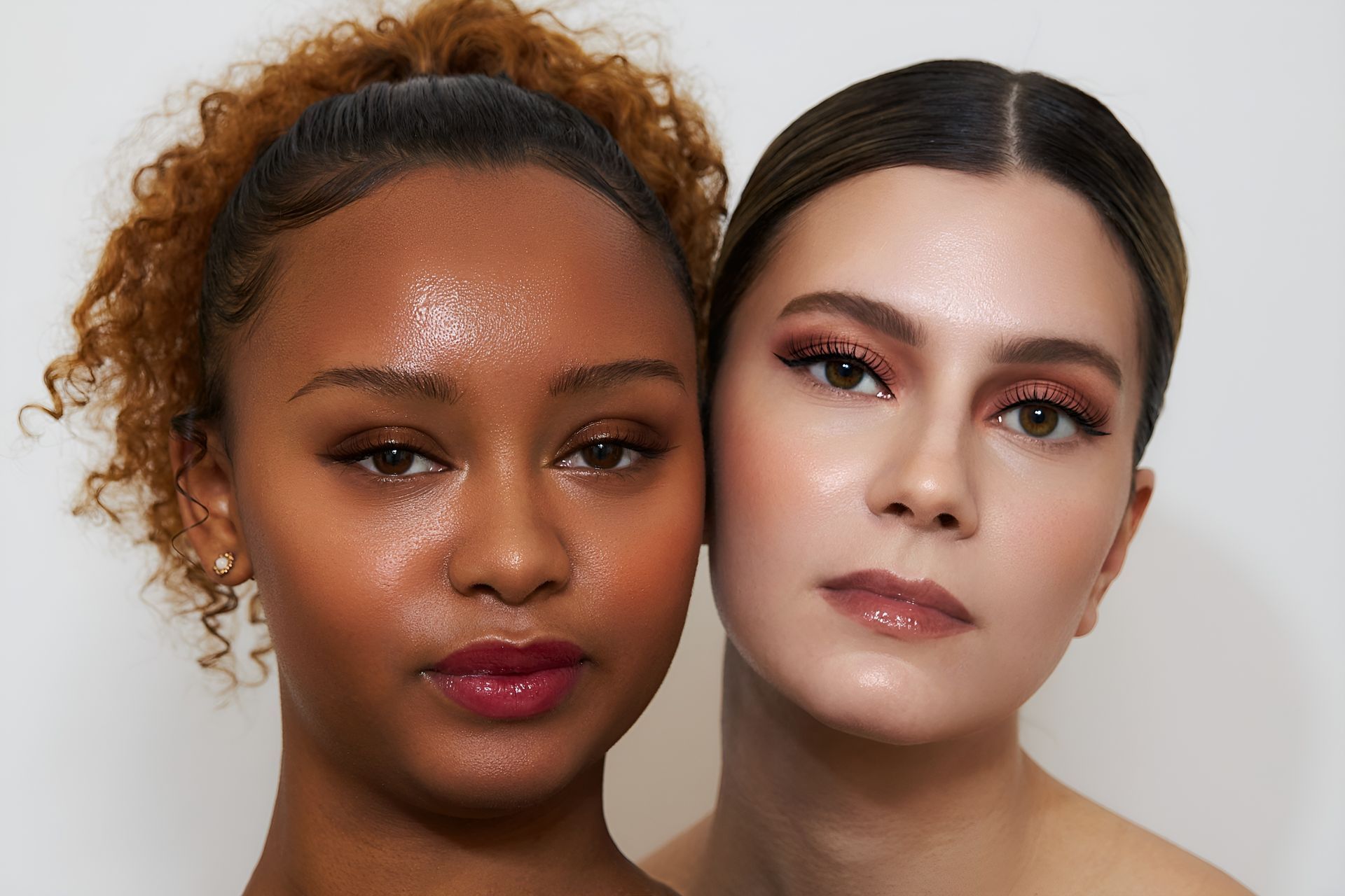 Zwei Frauen mit schimmernder Haut, eine dunkelhäutig, eine hellhäutig, blicken nach vorne. Beide tragen Make-up.